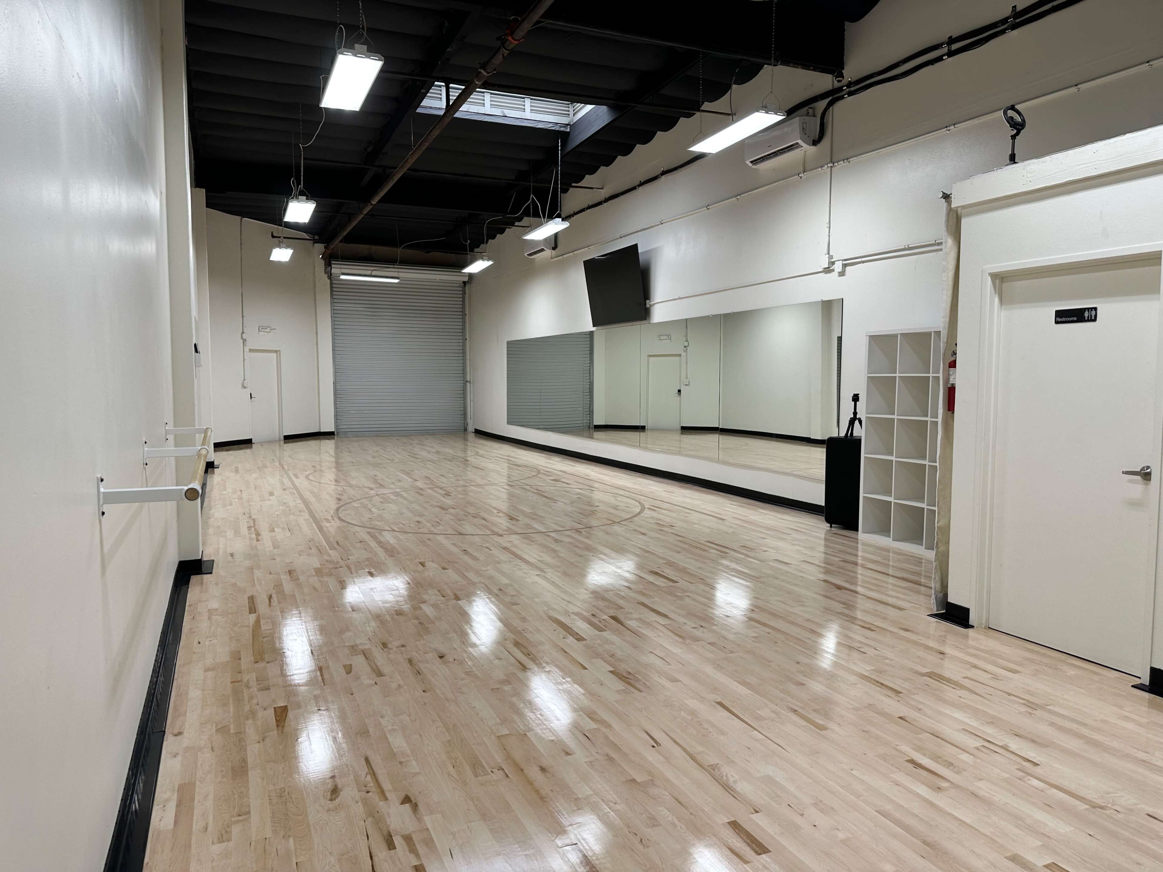 The image shows an empty dance studio with polished wooden flooring, a large mirror, and a closed garage door at one end.