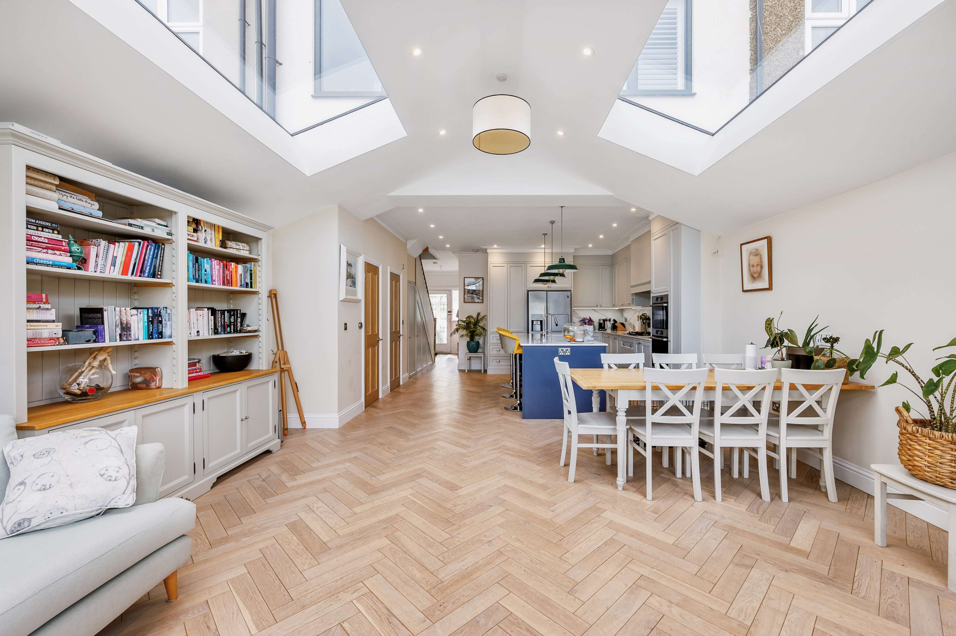 A bright, open-plan living space with a dining area, a kitchen, and a bookshelf, featuring large skylights that allow natural light to fill the room.