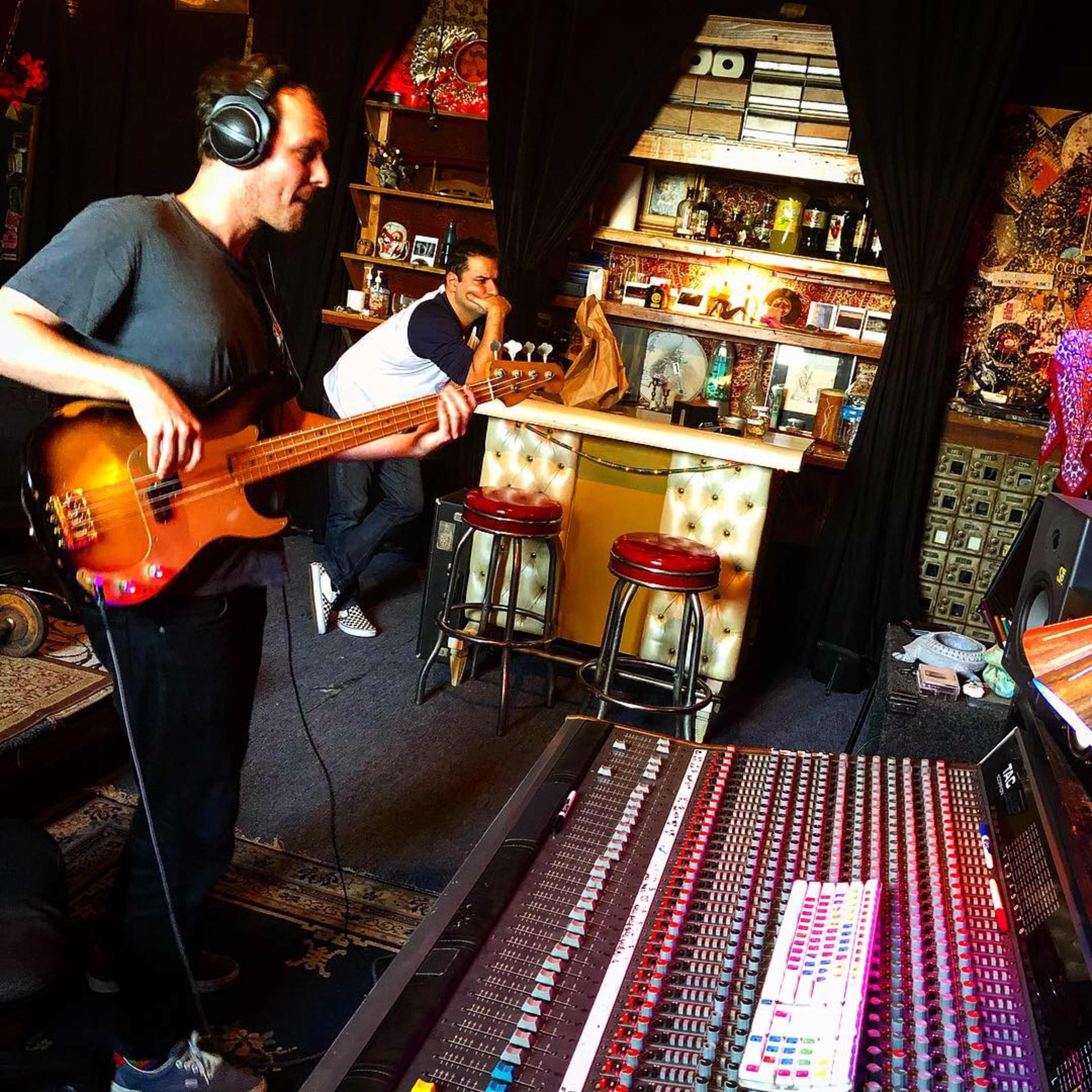 A man plays a bass guitar in a music studio while another man sits at a bar area in the background.