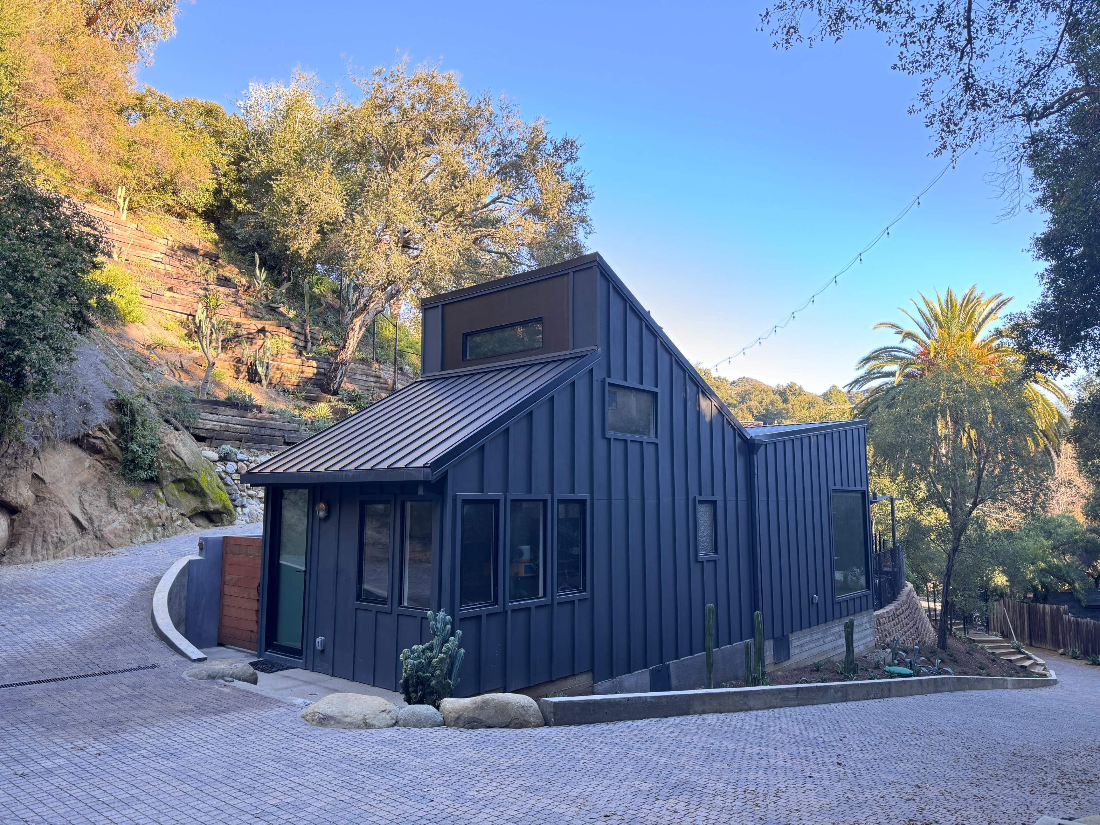 A modern, angular house with a metal exterior is situated on a winding driveway surrounded by trees and greenery.