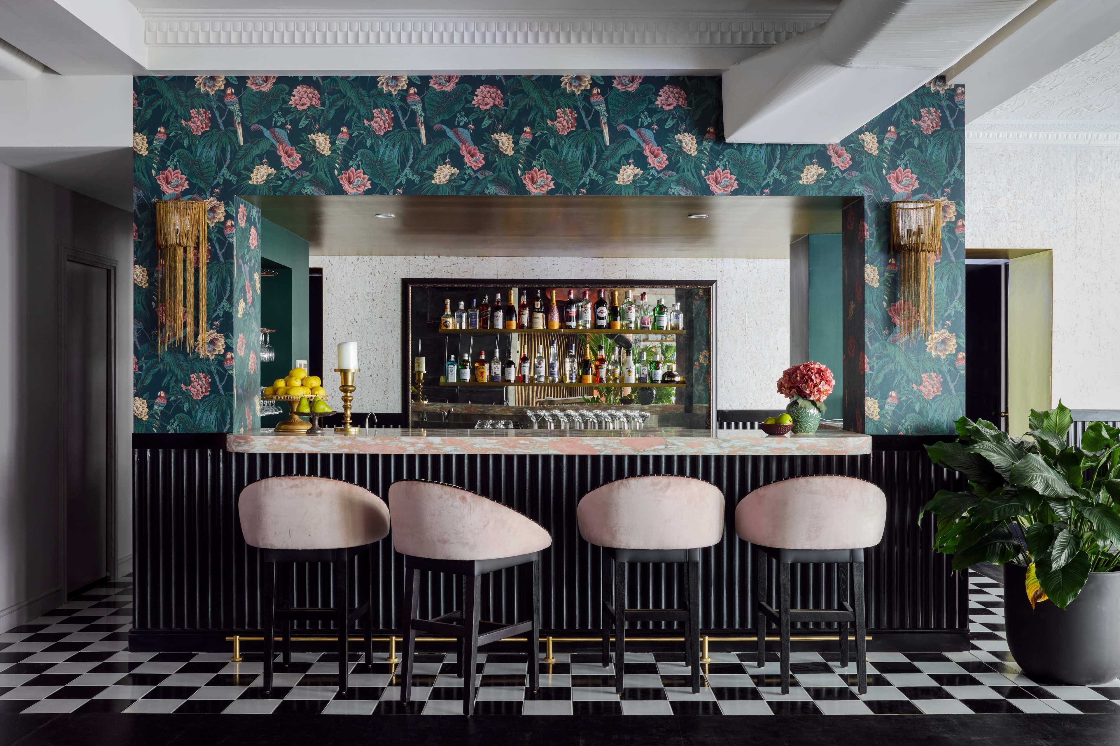 A stylish bar area with a floral wallpaper backdrop, a polished counter, and four upholstered stools in front.