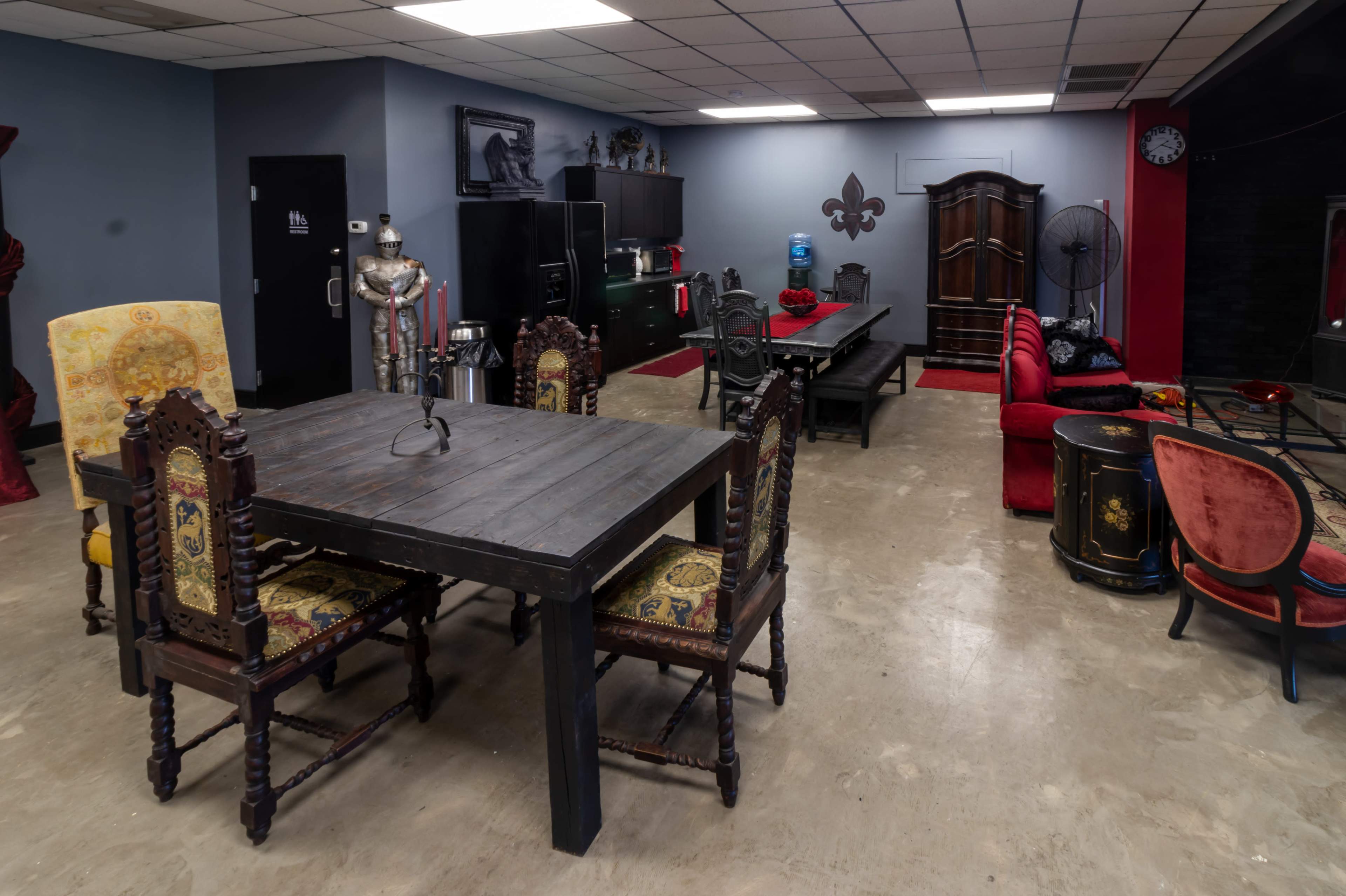 The image depicts a spacious room with a large wooden table surrounded by ornate chairs, a red couch, and various decorations including a suit of armor and a cabinet.