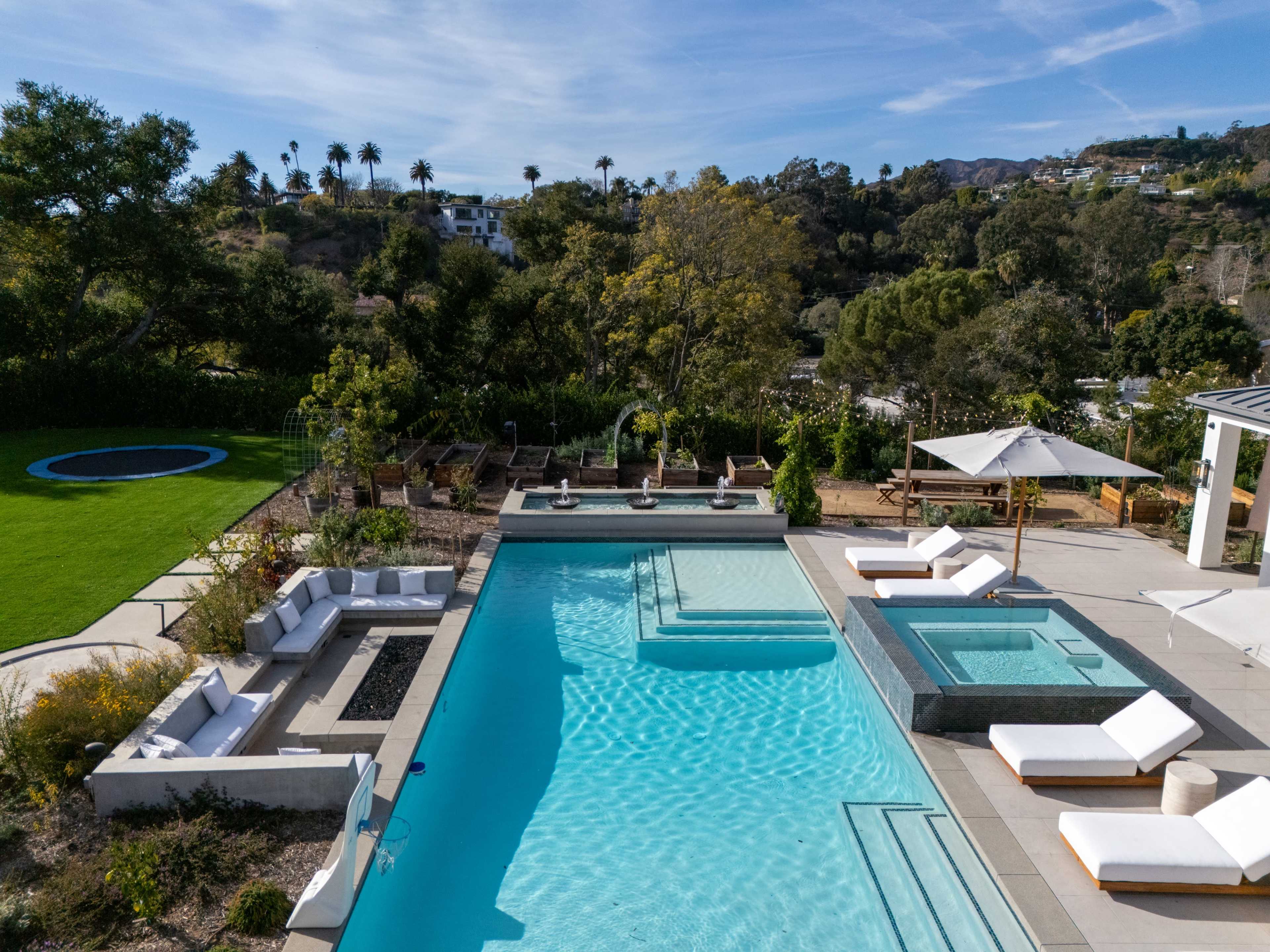 A modern outdoor space features a large swimming pool, a hot tub, lounge chairs, and landscaped greenery in the background.