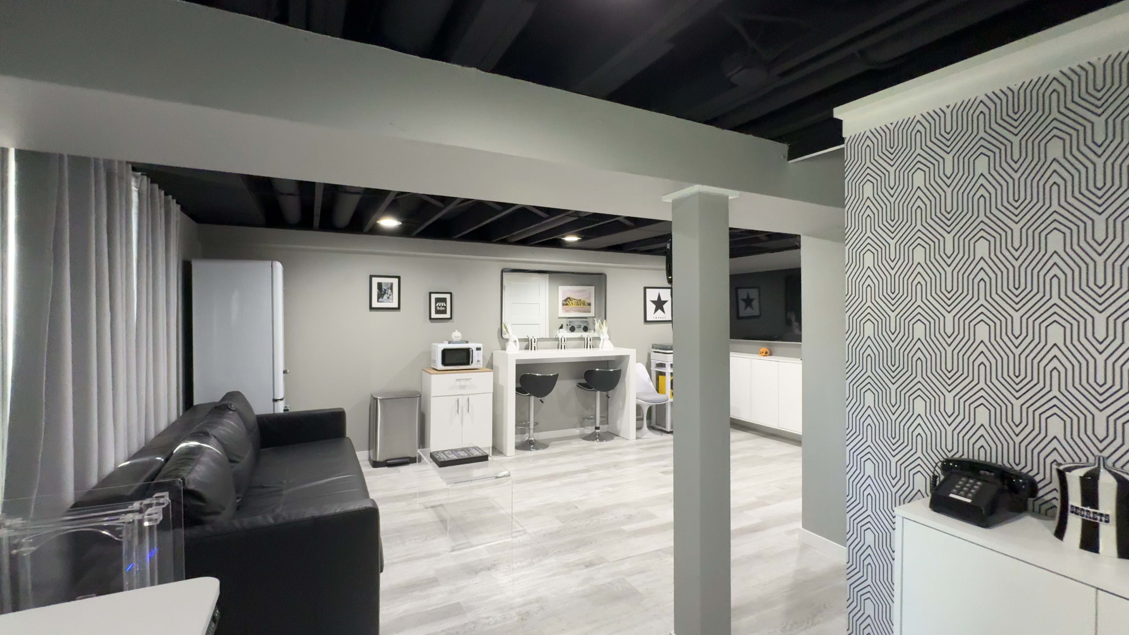 The image shows a modern, open-concept living space with a black leather couch, a kitchenette with bar stools, and white cabinets against a gray wall.
