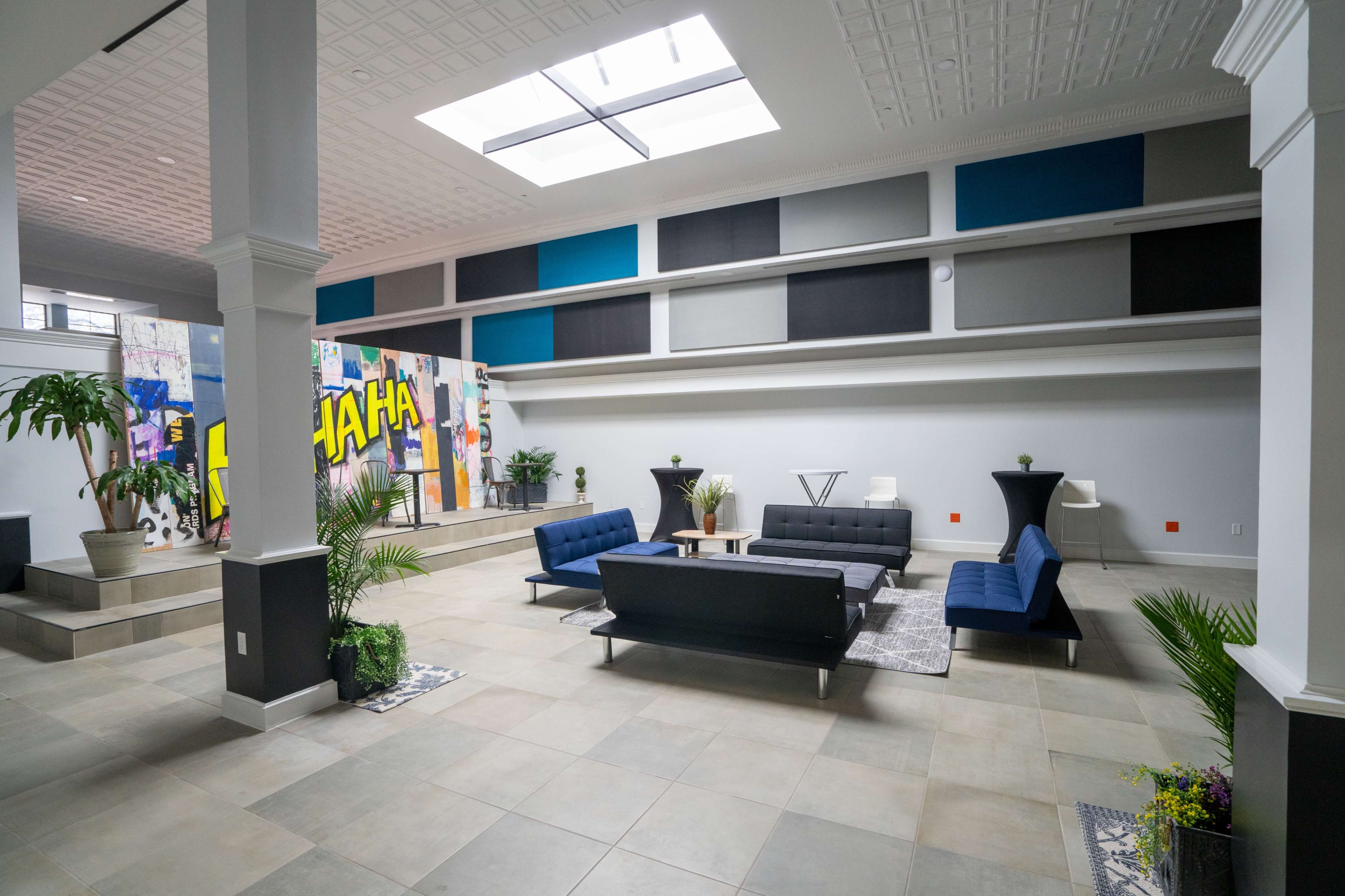 A spacious room with a modern seating area that includes two blue sofas, a coffee table, and decorative plants, along with a colorful mural on one wall.