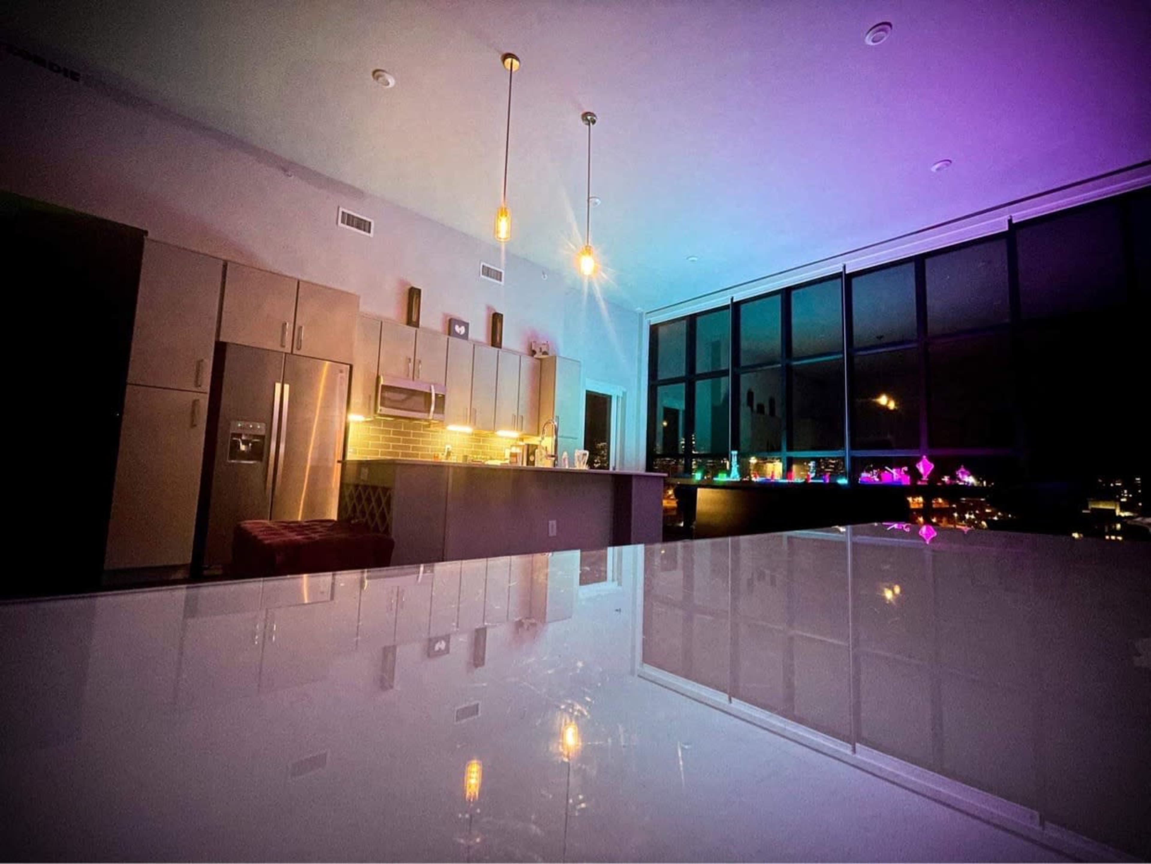A modern kitchen with sleek cabinetry and lighting that casts colorful hues across the walls and a glossy counter.