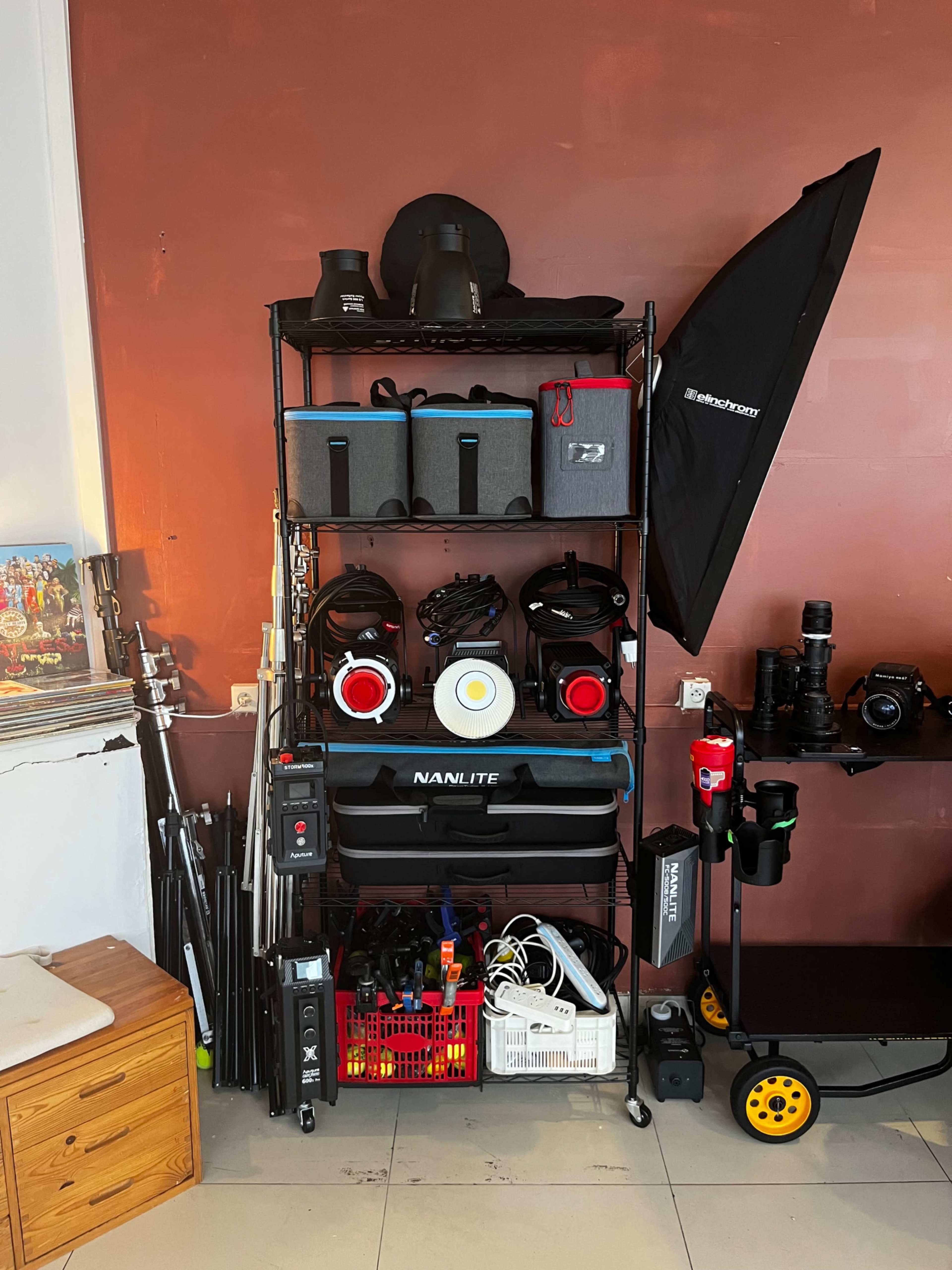 The image shows a shelf organized with various photography and lighting equipment, including boxes, light stands, and camera gear, against a brown wall.