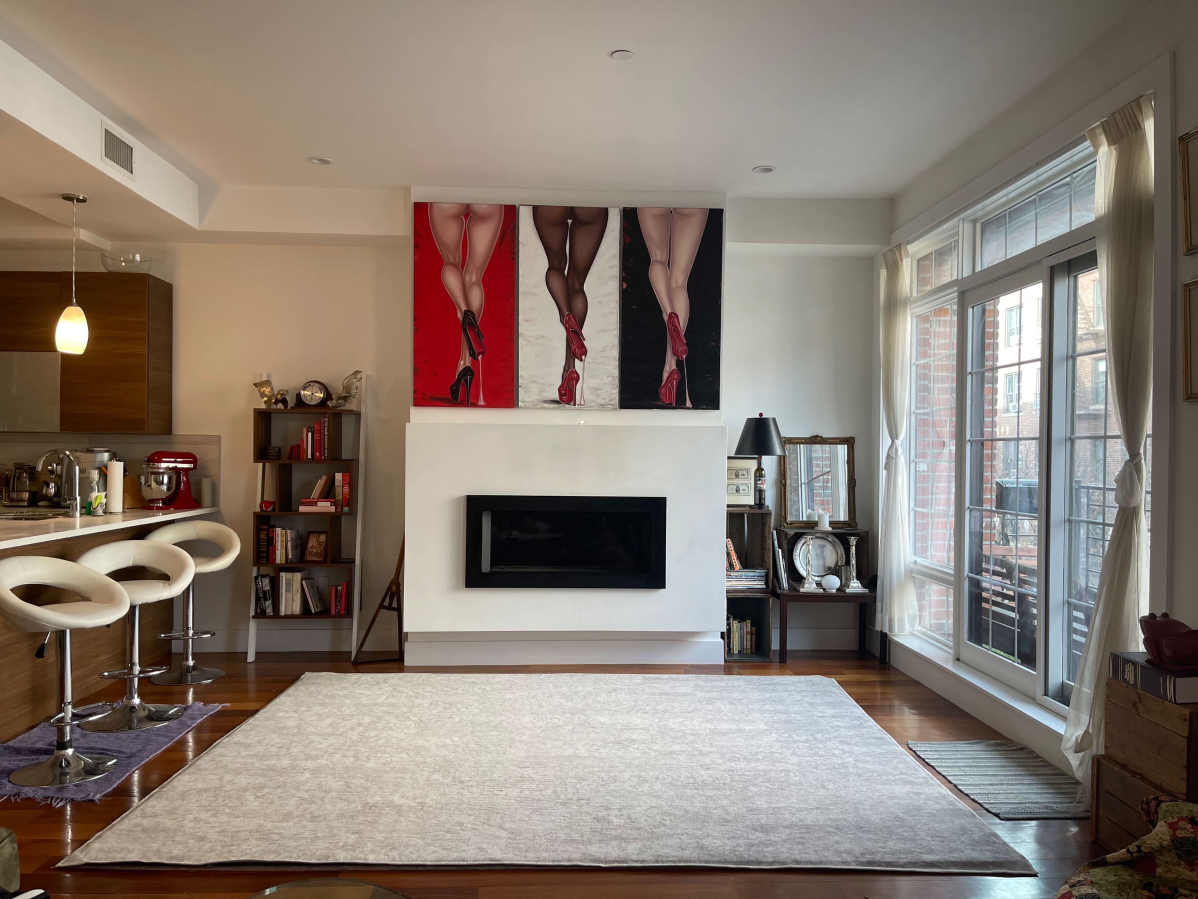 A modern living room features a large artwork of legs in high heels above a sleek fireplace, surrounded by warm wood flooring and contemporary furniture.