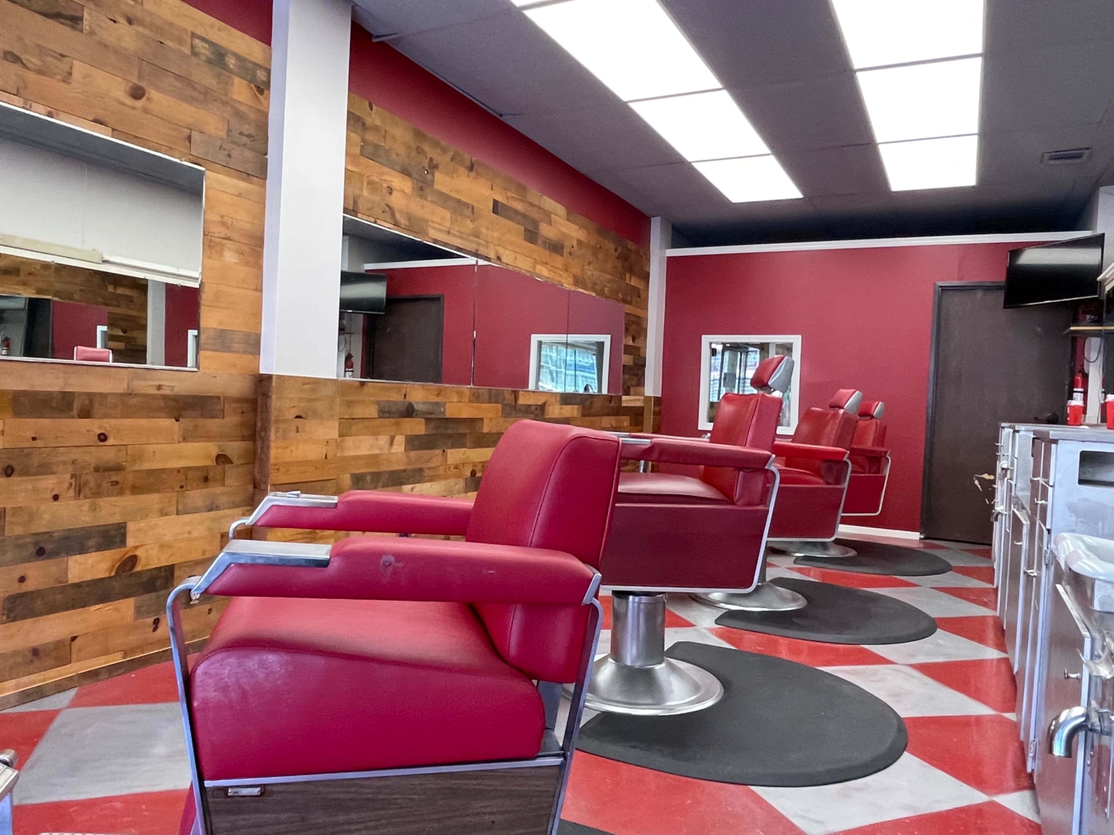 A barber shop interior featuring red-and-white checkered flooring, several red barber chairs, and wooden wall paneling with mirrors.