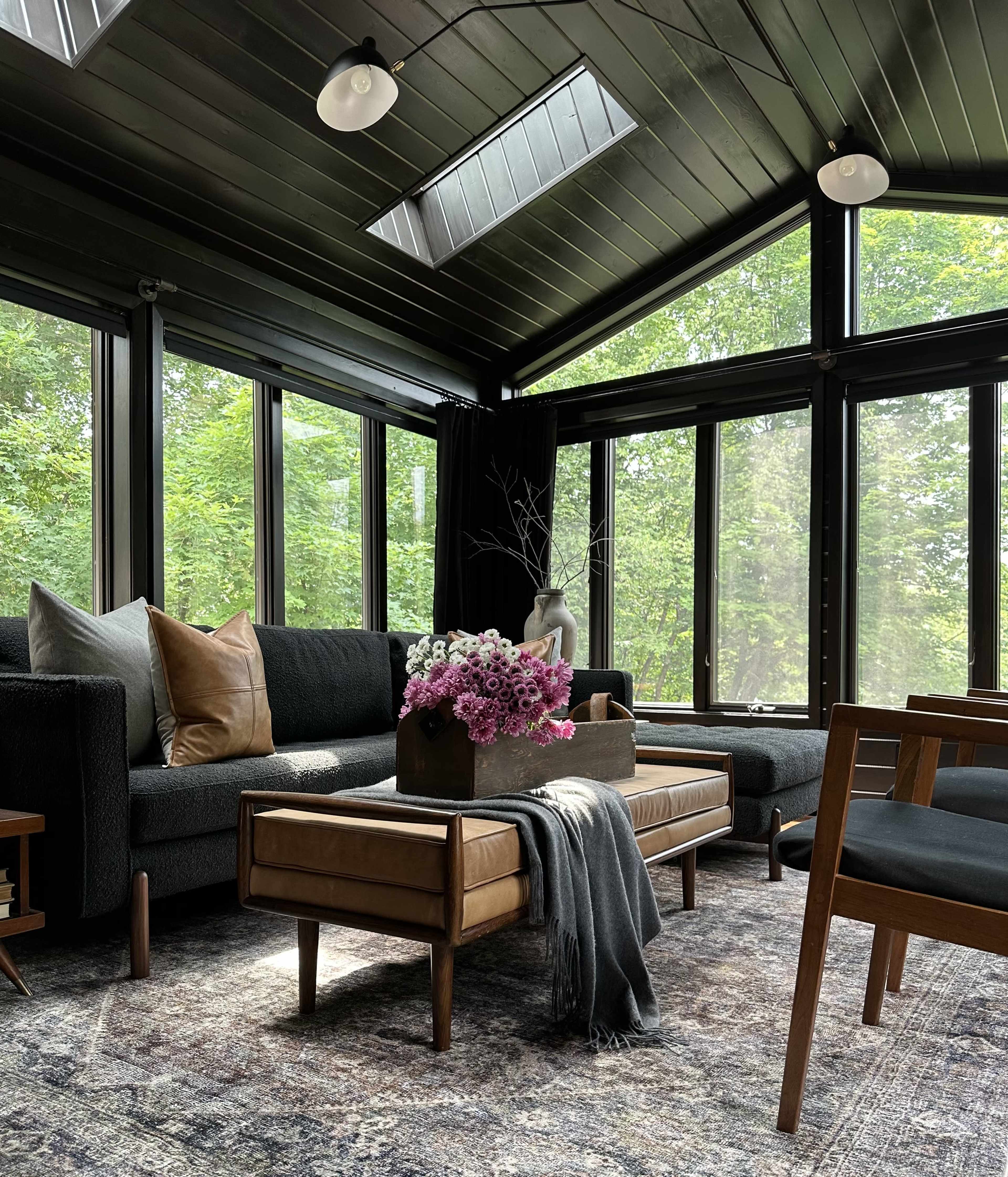 The interior of a sunlit living room features dark walls, large windows, a dark sectional sofa, a wooden coffee table with flowers, and a patterned rug.