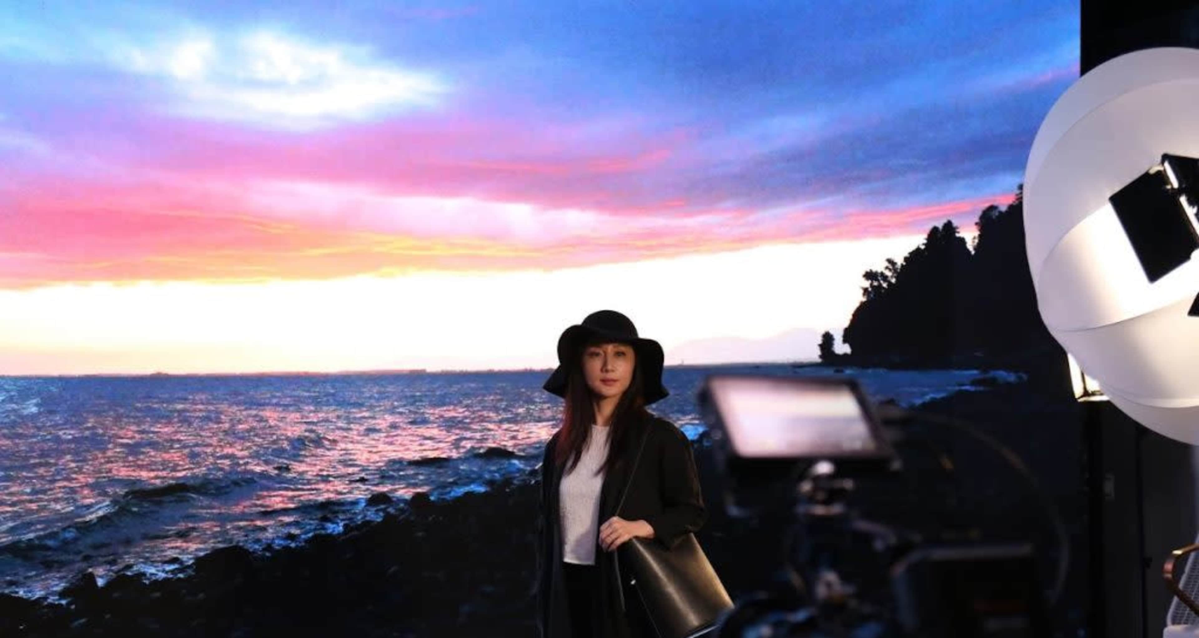A woman in a black outfit and hat poses in front of a colorful sunset backdrop near the ocean.