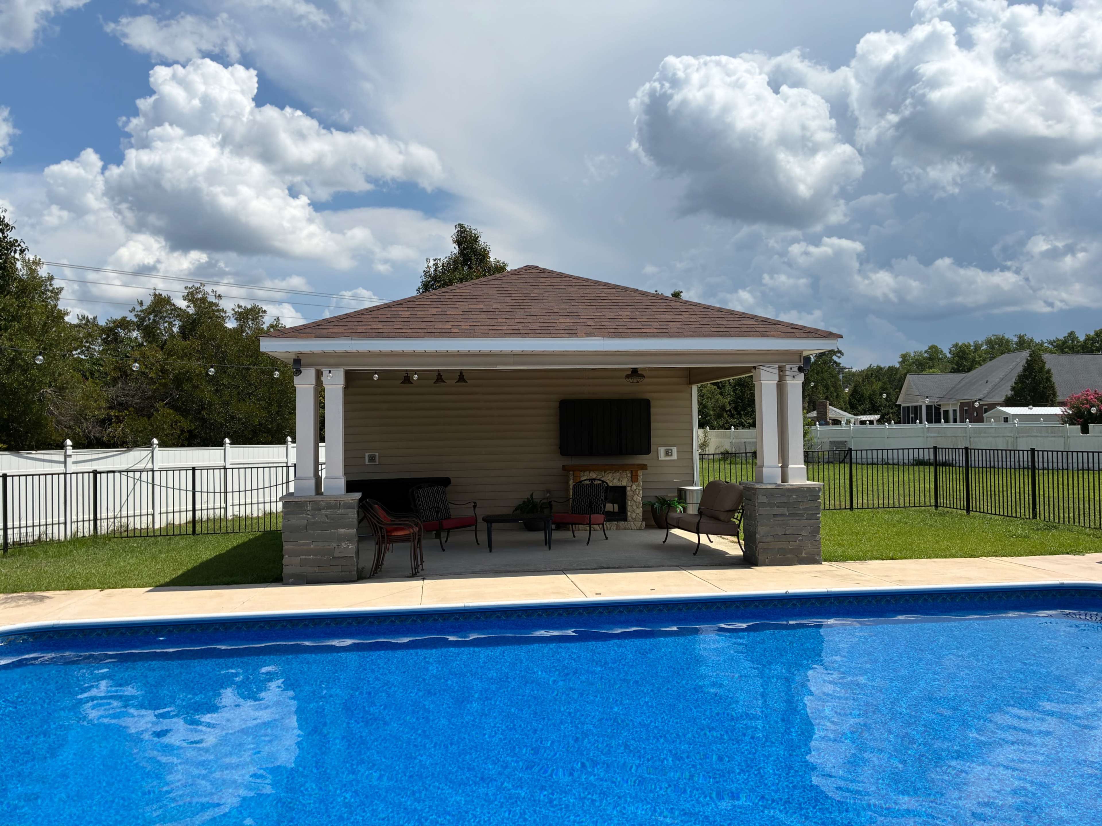 A covered patio with seating and a television is situated beside a swimming pool in a landscaped backyard under a cloudy sky.