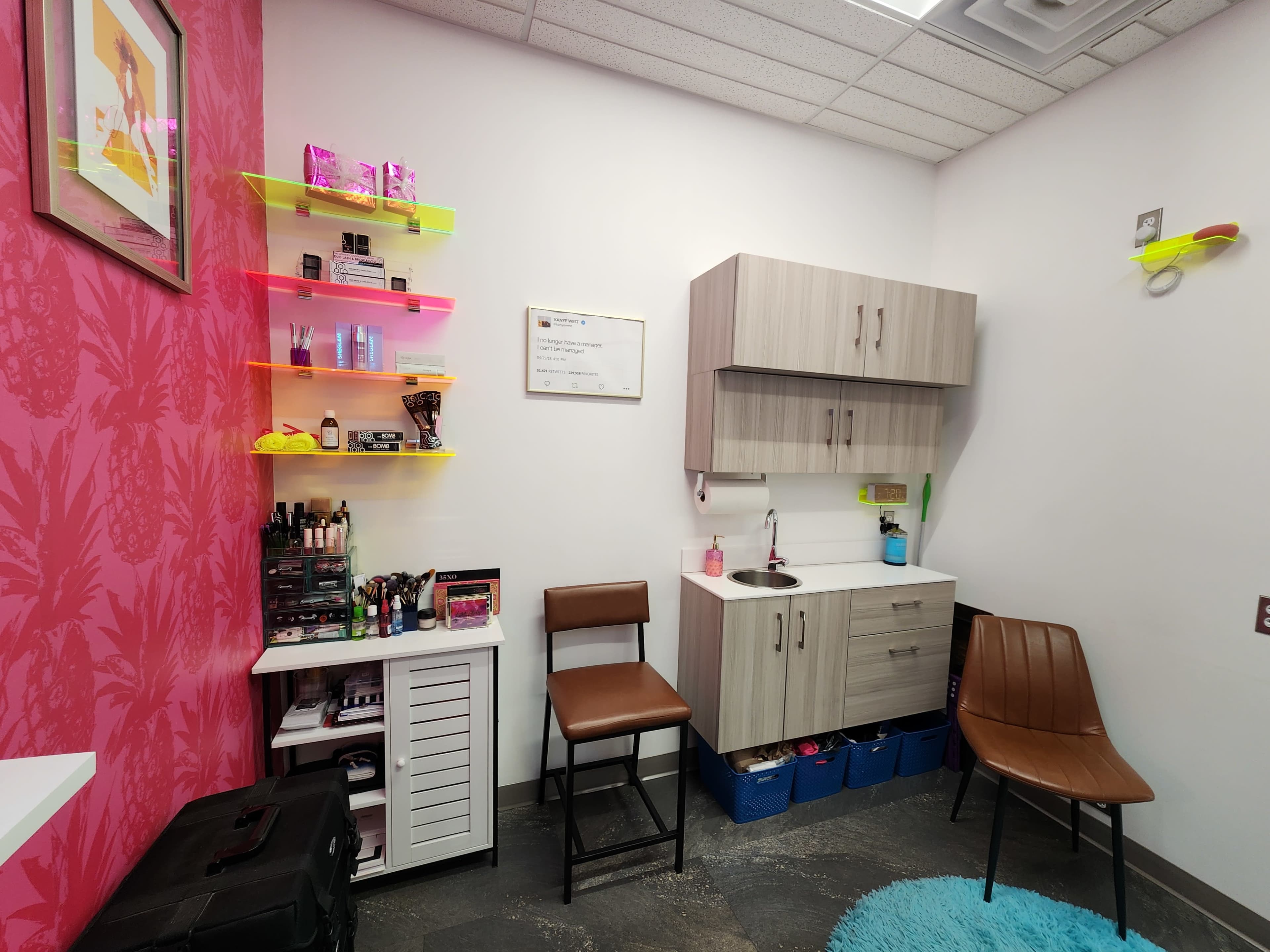 The image shows a brightly colored room with pink walls, shelves displaying various beauty products, and seating along with a vanity area that includes a sink and storage cabinets.