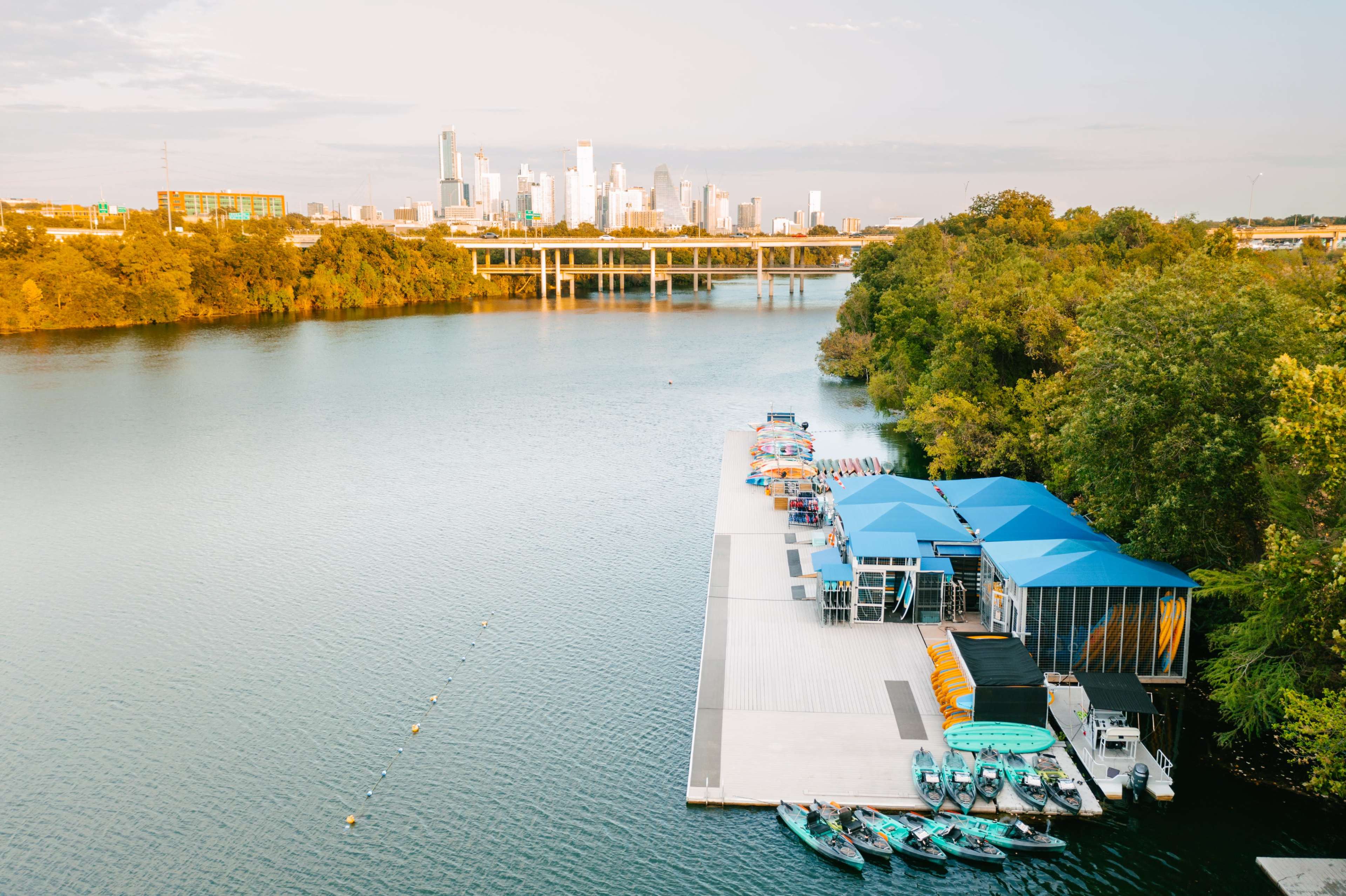 A dock with kayaks and canoes is positioned along a river, with a city skyline visible in the background.