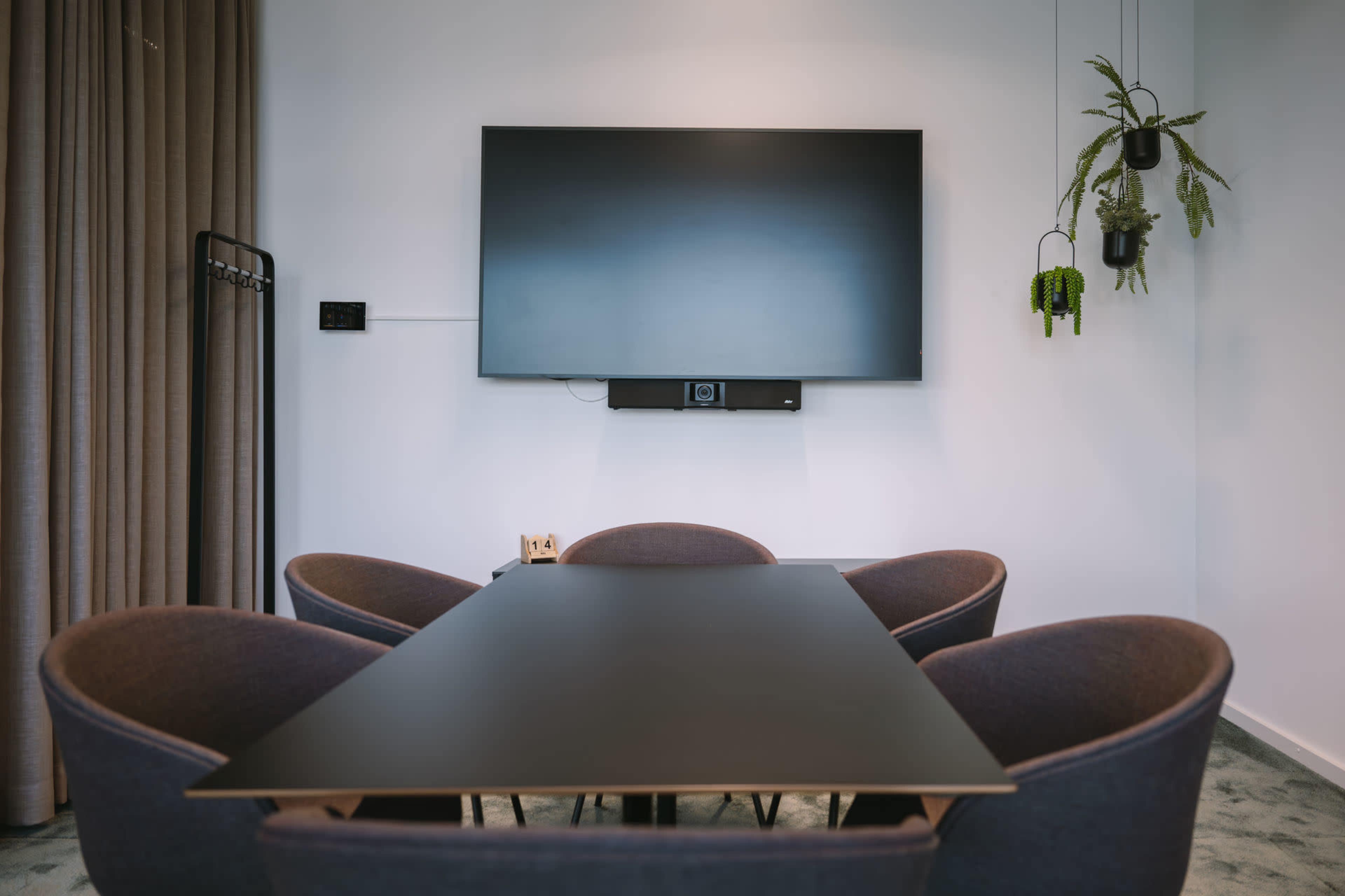 A conference room features a long table surrounded by four chairs, with a large screen mounted on the wall and greenery hanging nearby.