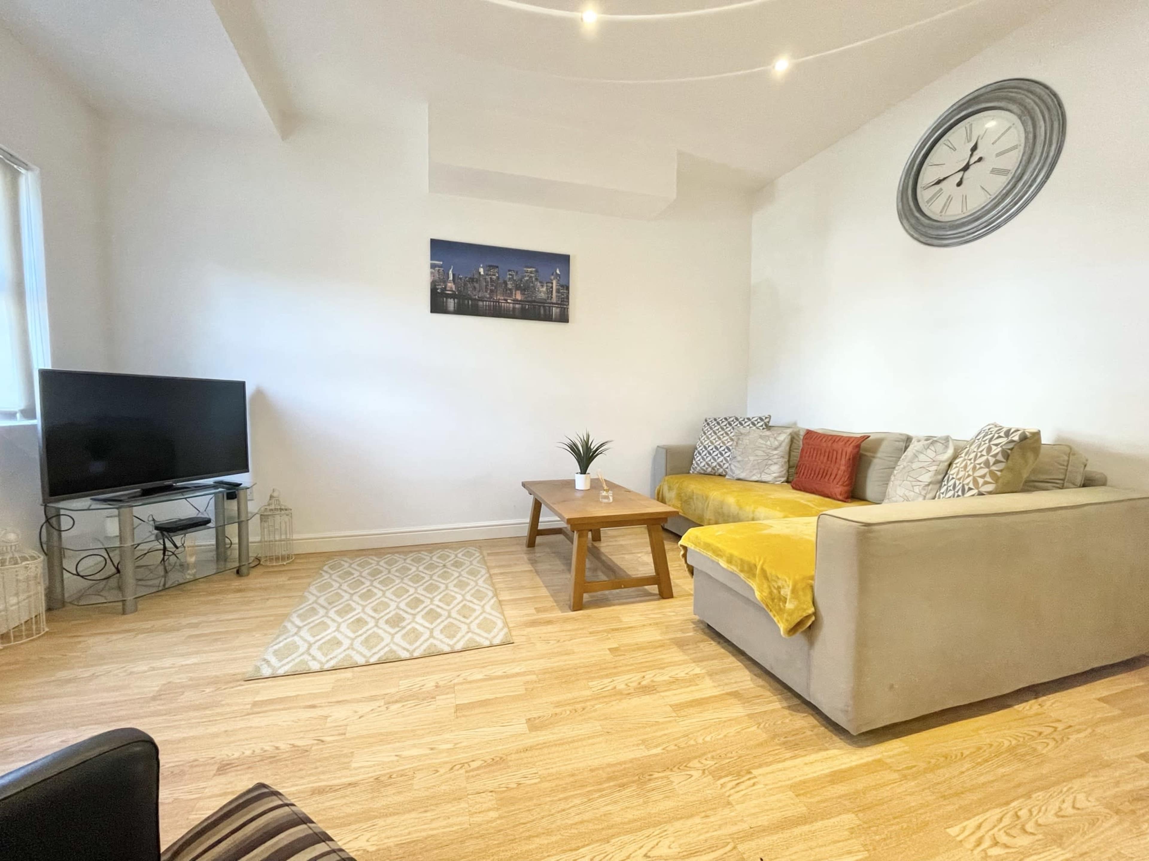 A living room with a television, a coffee table, a sectional sofa, a wall clock, and light wooden flooring.