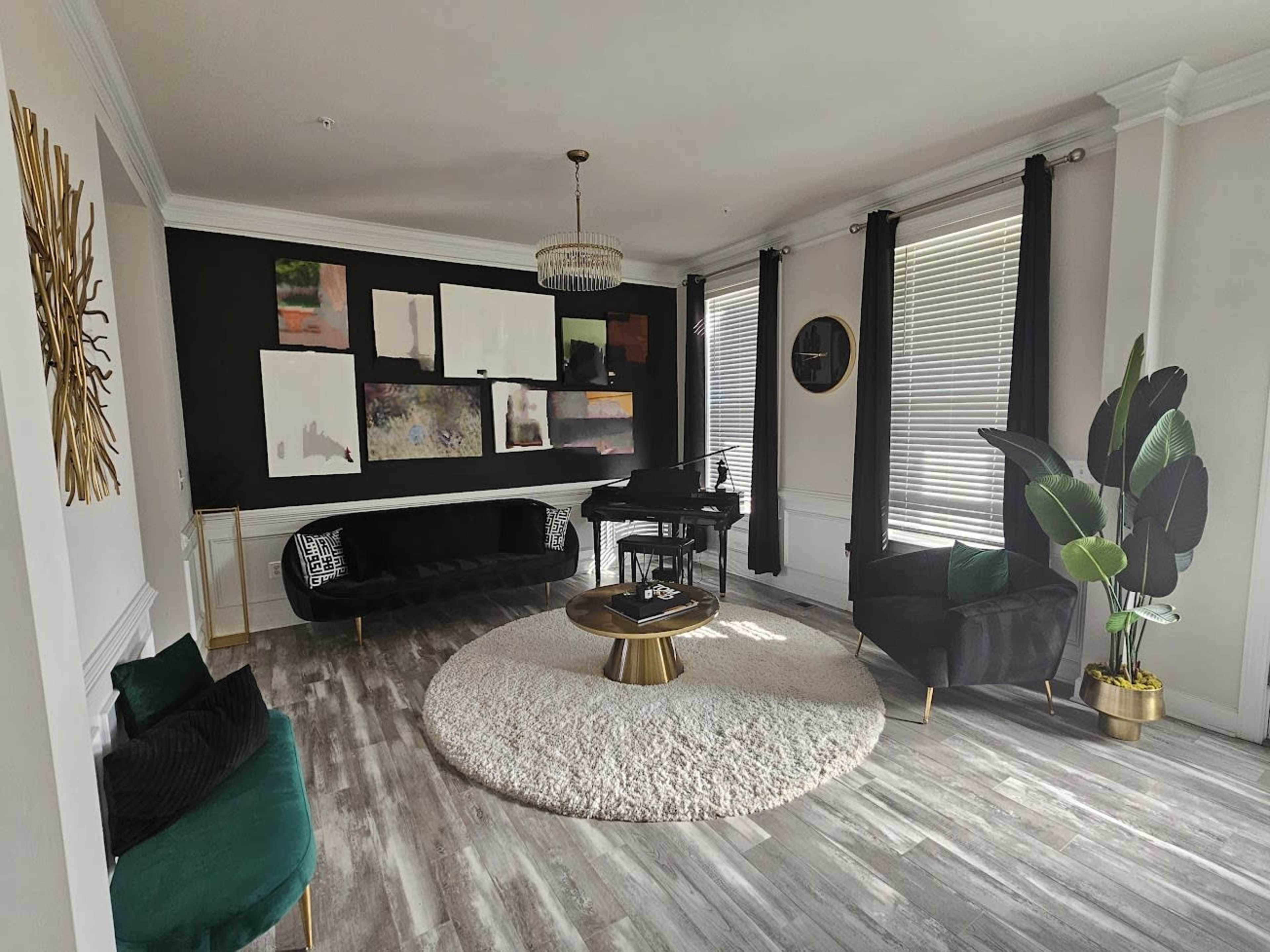 The image shows a modern living room with a grand piano, green velvet furniture, a round white rug, and large windows with black curtains.