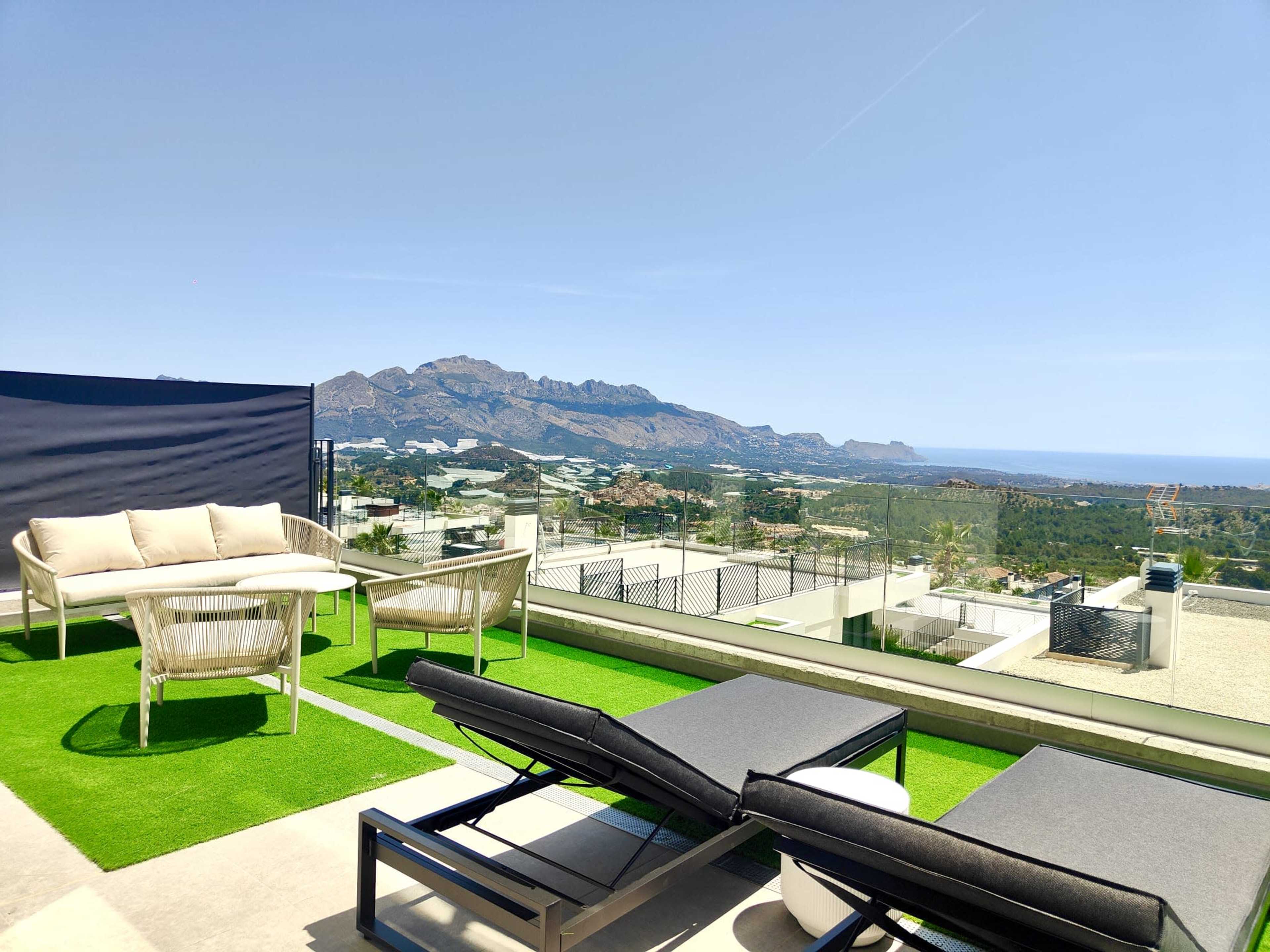 A rooftop terrace features modern lounge furniture with a view of mountains and the sea in the background.