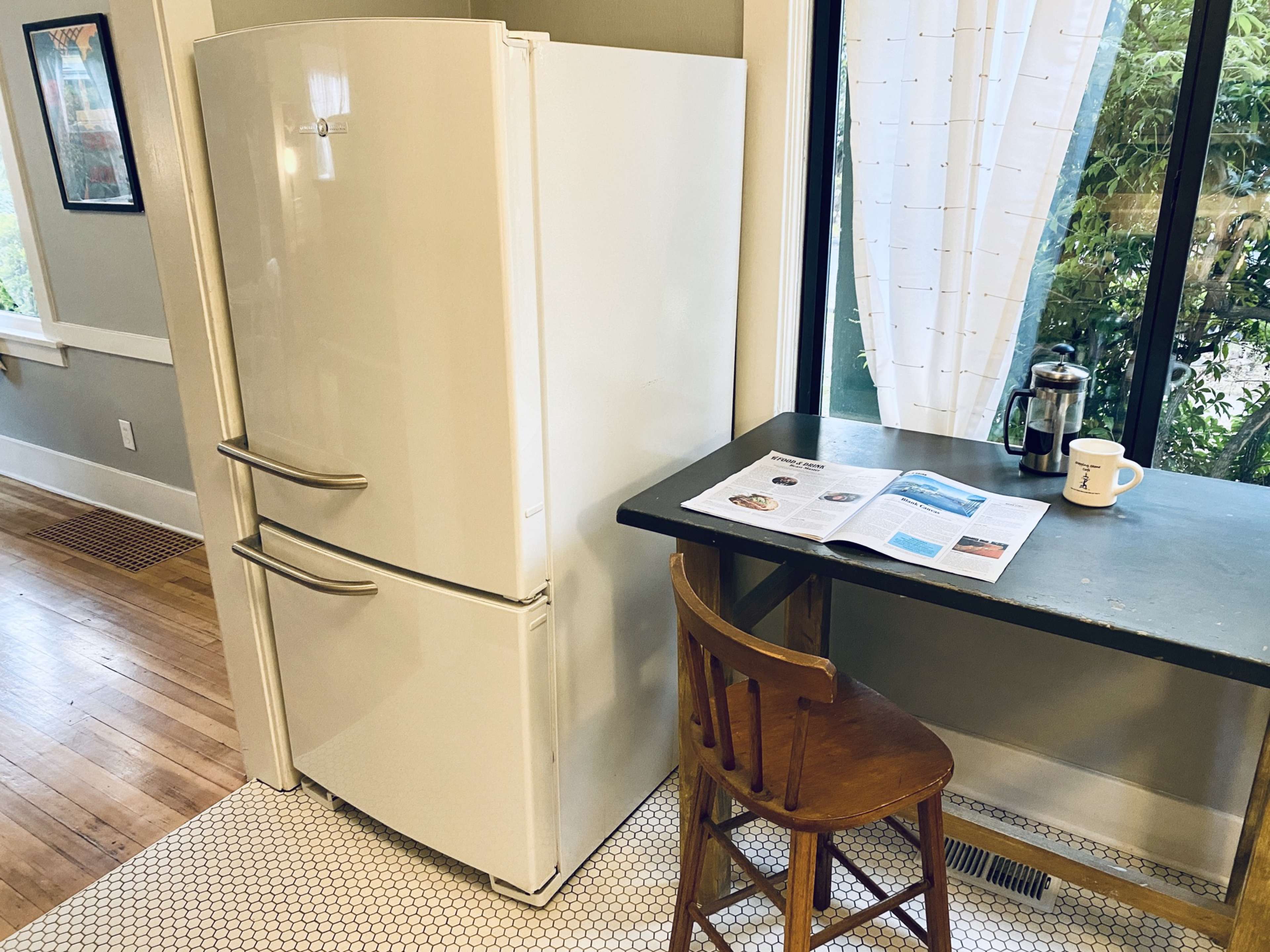 A white refrigerator stands next to a small dining table with a magazine and a coffee cup on it, set against a backdrop of a window and greenery outside.