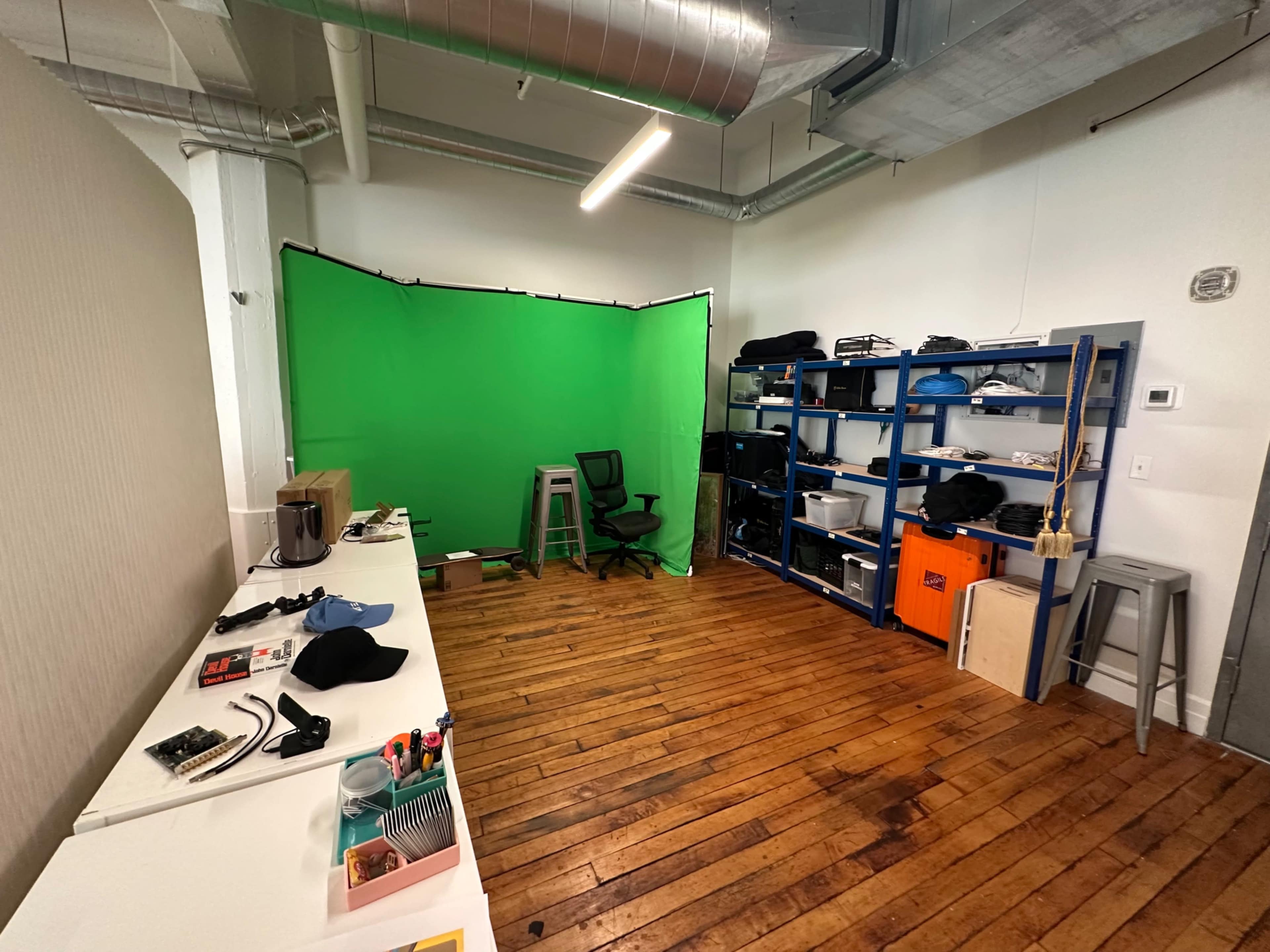 The image shows a spacious room with wooden floors, featuring a green backdrop, a chair, a shelving unit filled with various items, and a long table with tools and equipment.