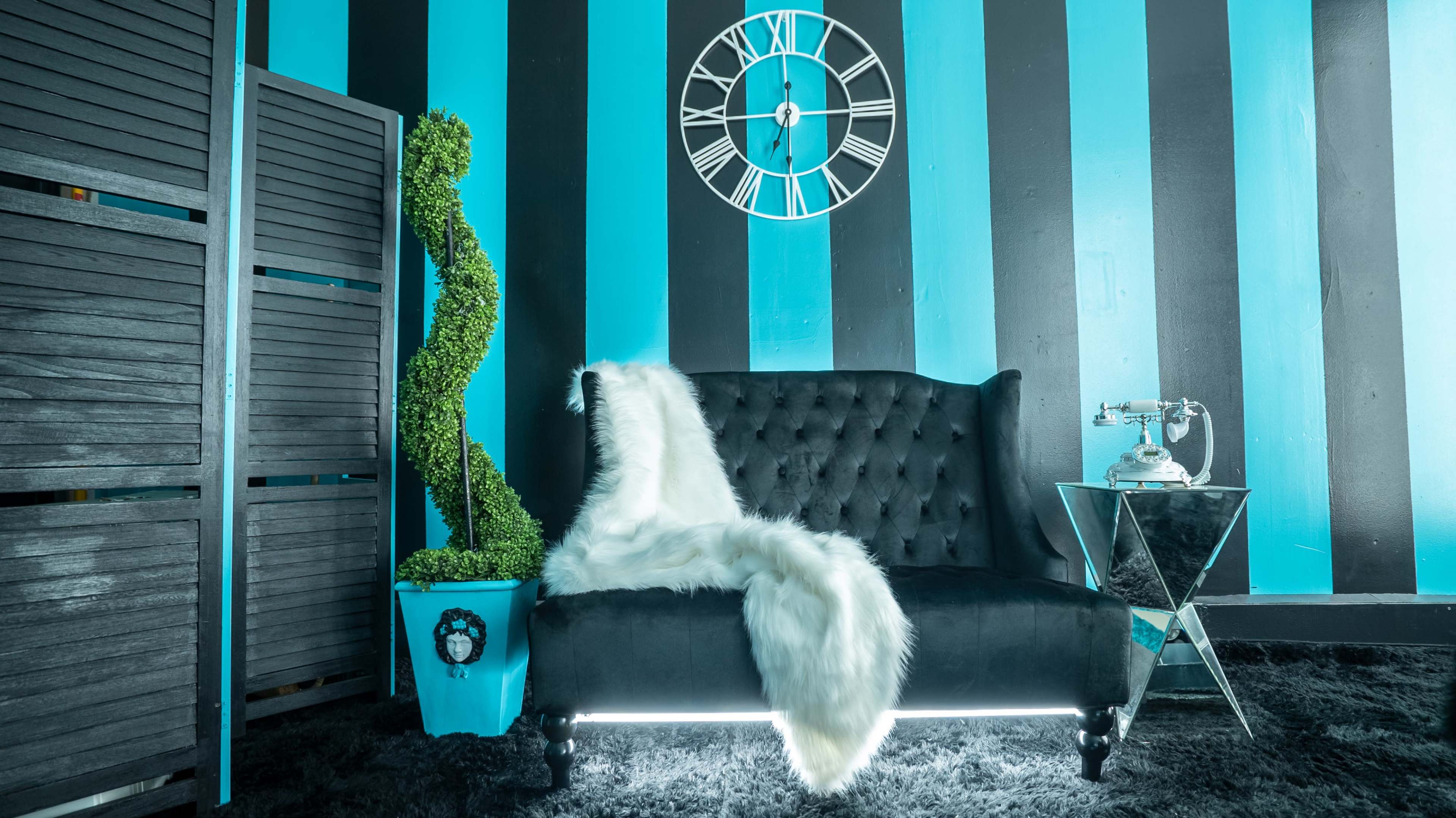 The image shows a black tufted sofa adorned with a white fur throw, positioned against blue and black striped walls, accompanied by a clock, a decorative plant, and a glass side table with a vintage telephone.