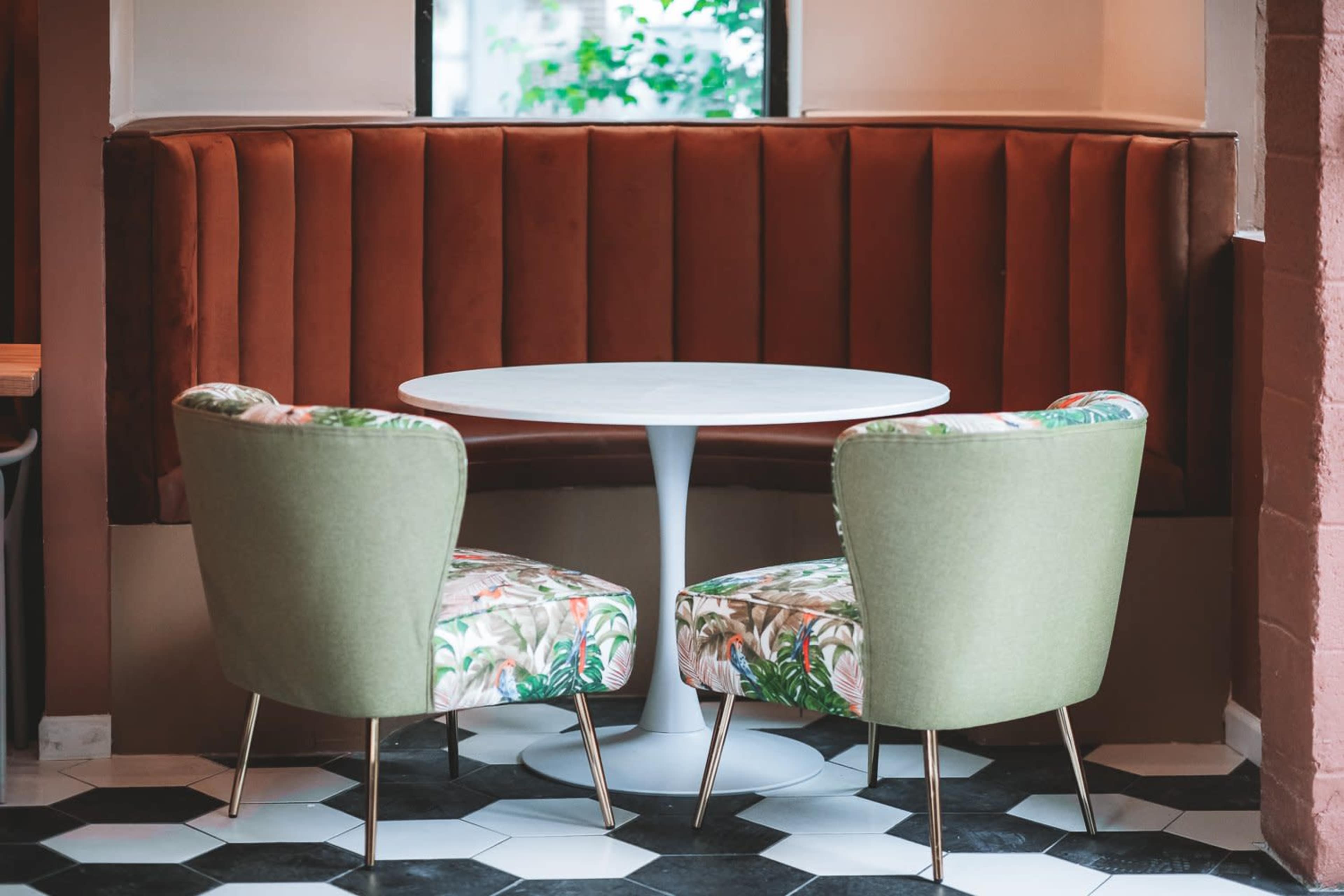 A round white table is set between two upholstered chairs with floral patterns against a backdrop of a tufted orange banquette.