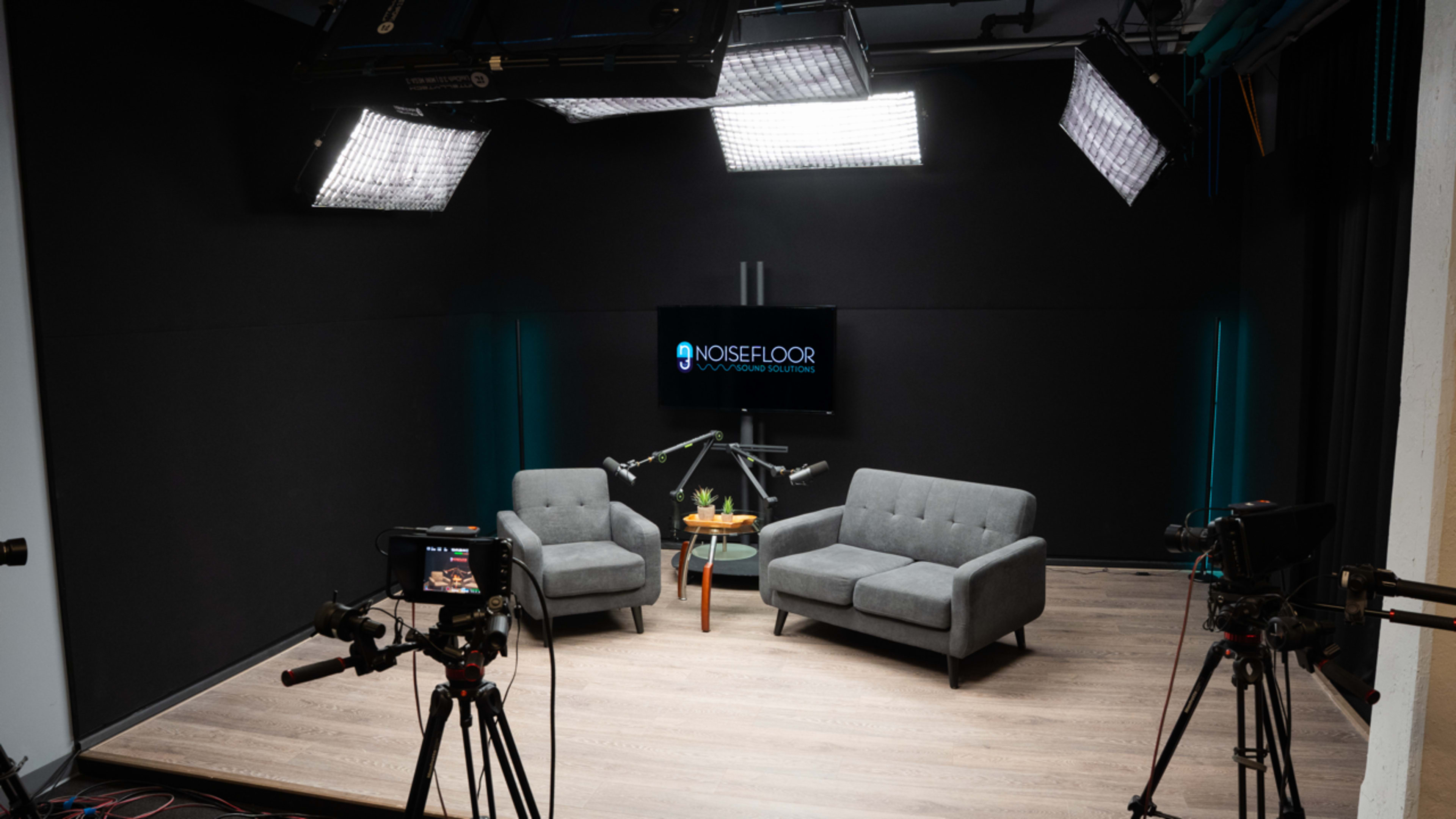 The image shows a modern studio setup with two gray chairs, a wooden table with a plant, and cameras positioned around for filming.