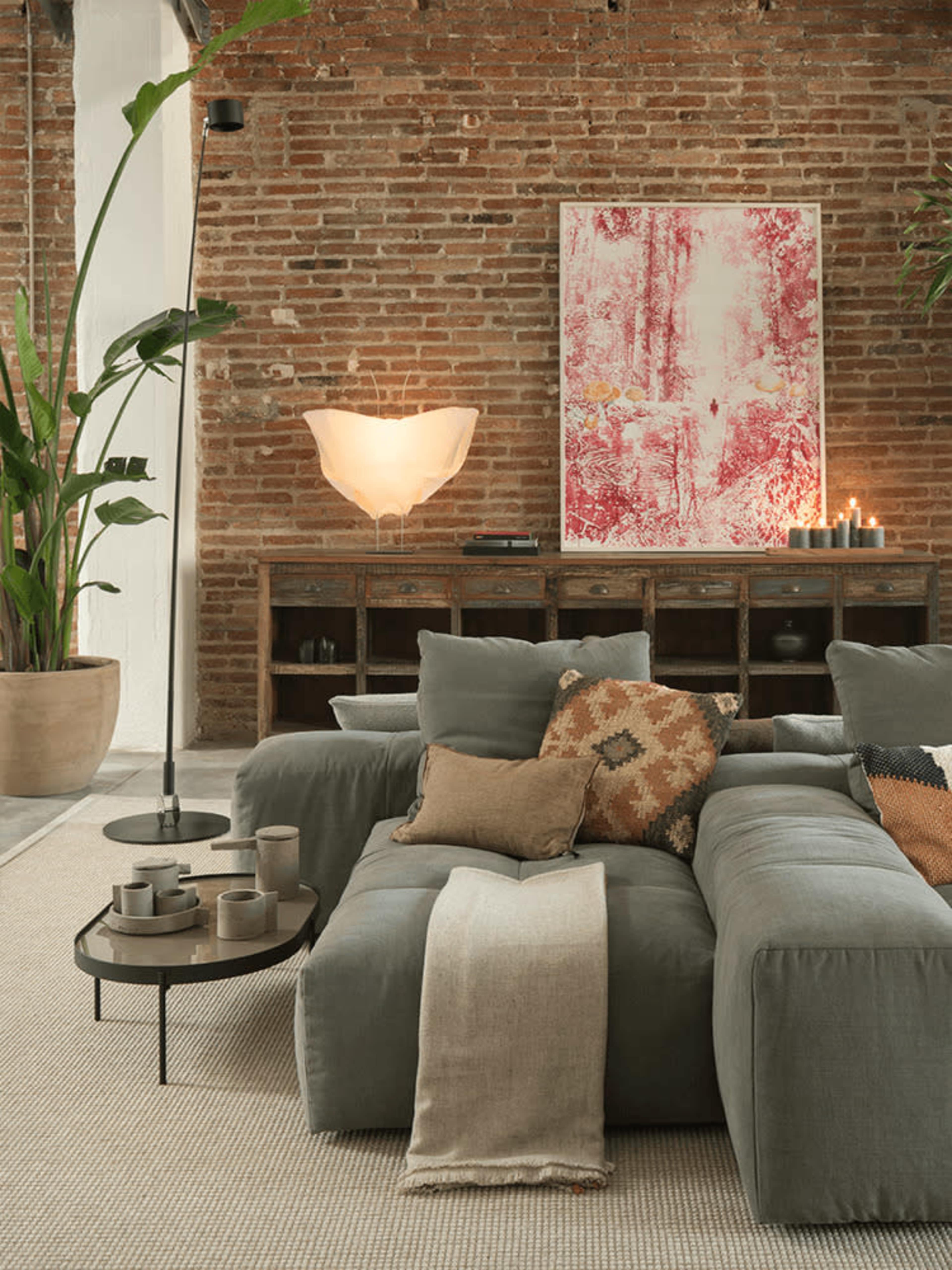 The image features a stylish living room with a large gray sofa, a wooden console, a floor lamp, and decorative items set against a backdrop of exposed red brick walls.