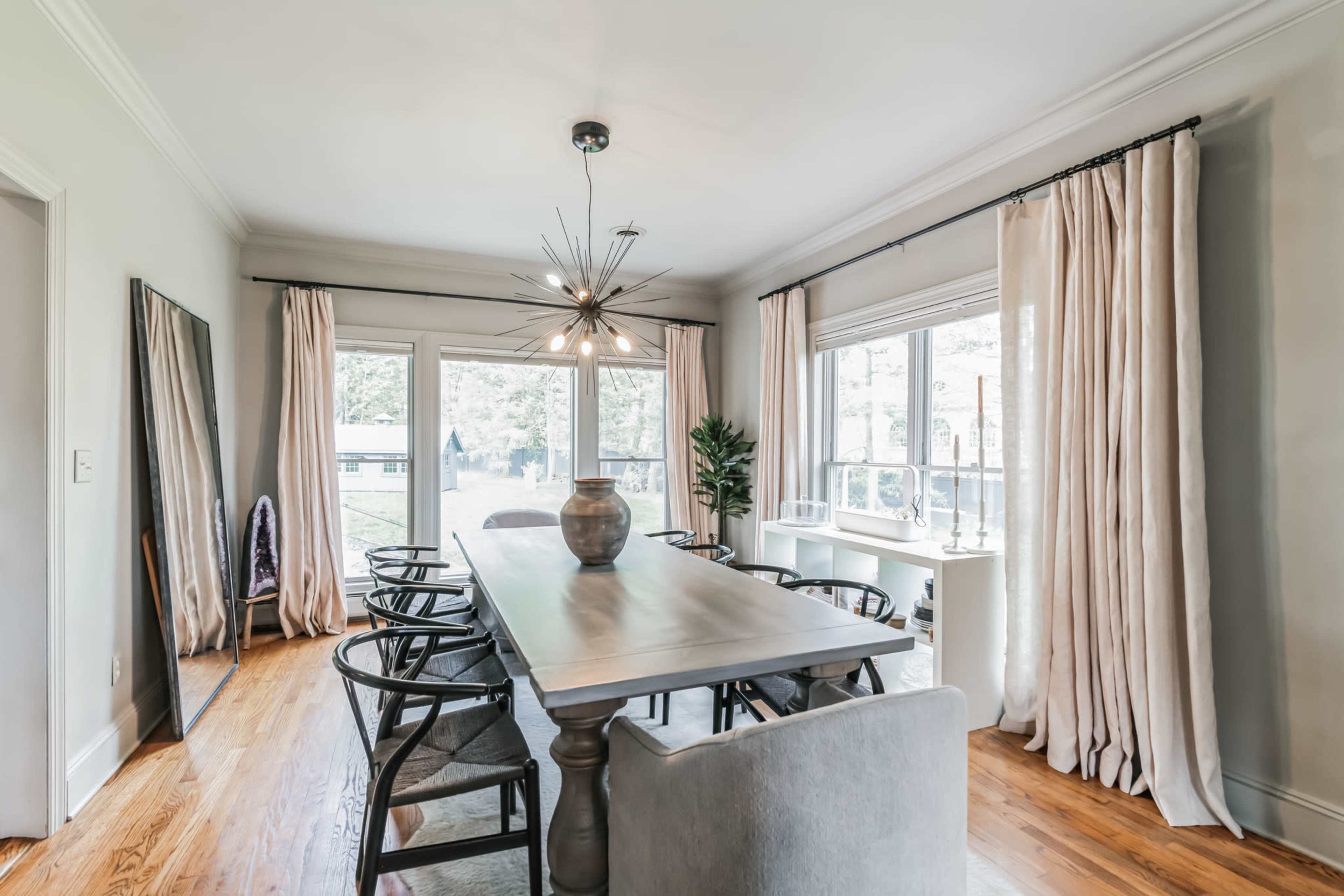 A spacious dining room with a long wooden table, surrounded by black chairs, large windows, and curtains, giving a view of the outdoors.