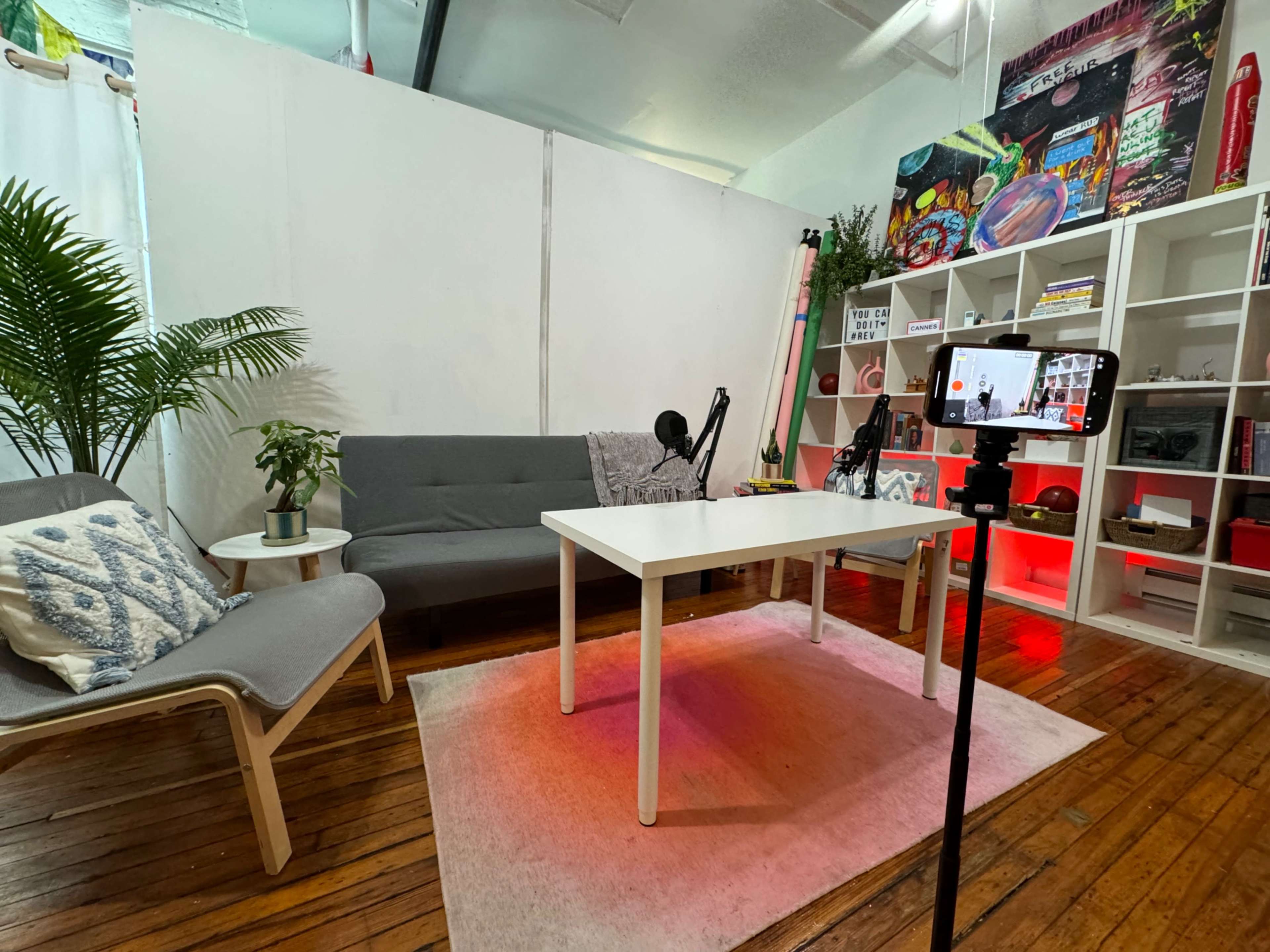 The image shows a modern interior setup featuring a white table, two couches, a plant, and shelving with colorful decor and lighting.