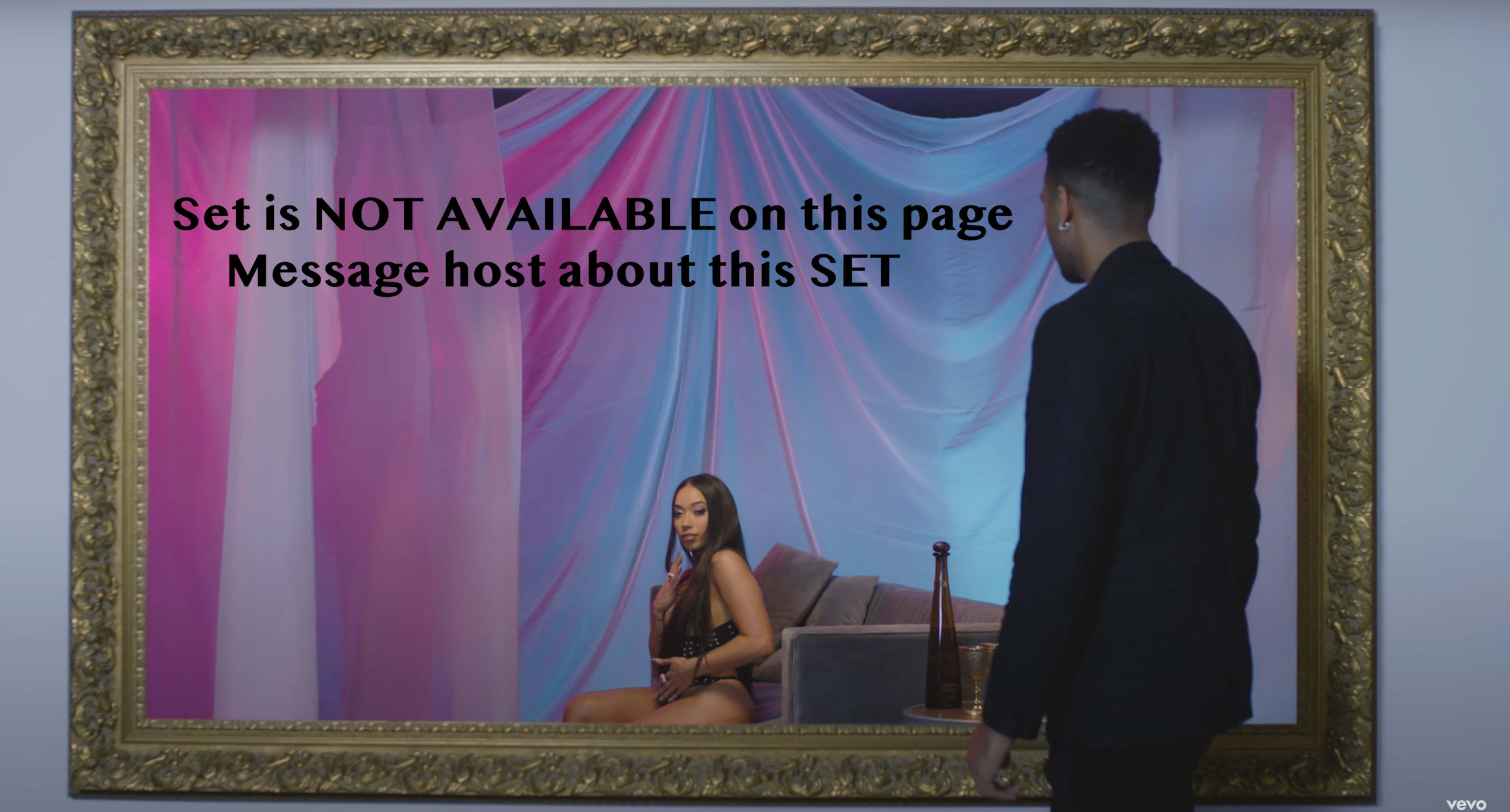 A man stands in front of a large, framed mirror, reflecting a woman seated on a couch against a backdrop of draped fabric in varying colors.