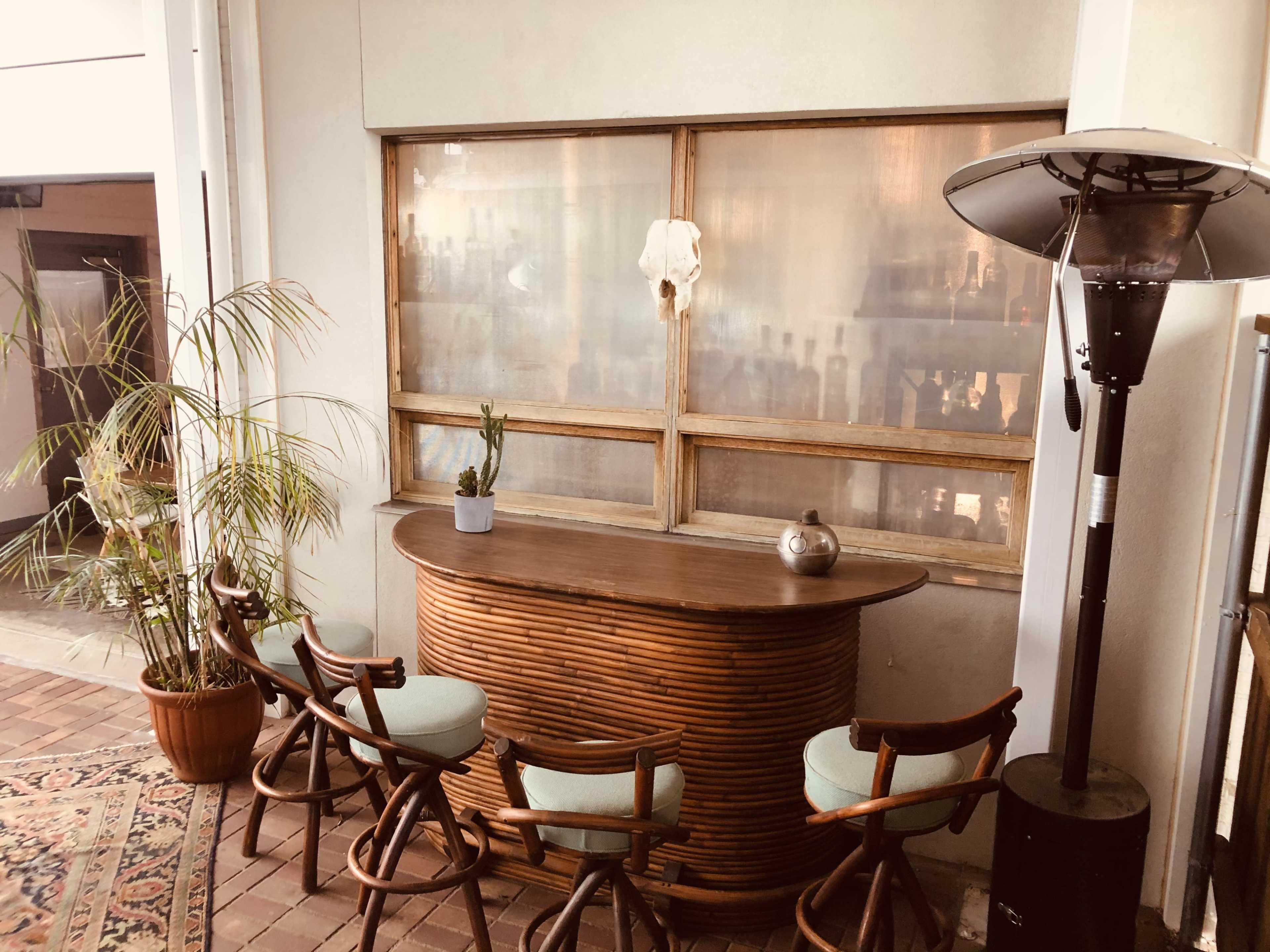 A curved wooden bar is positioned against a window with a potted plant nearby and several wooden chairs arranged around it.