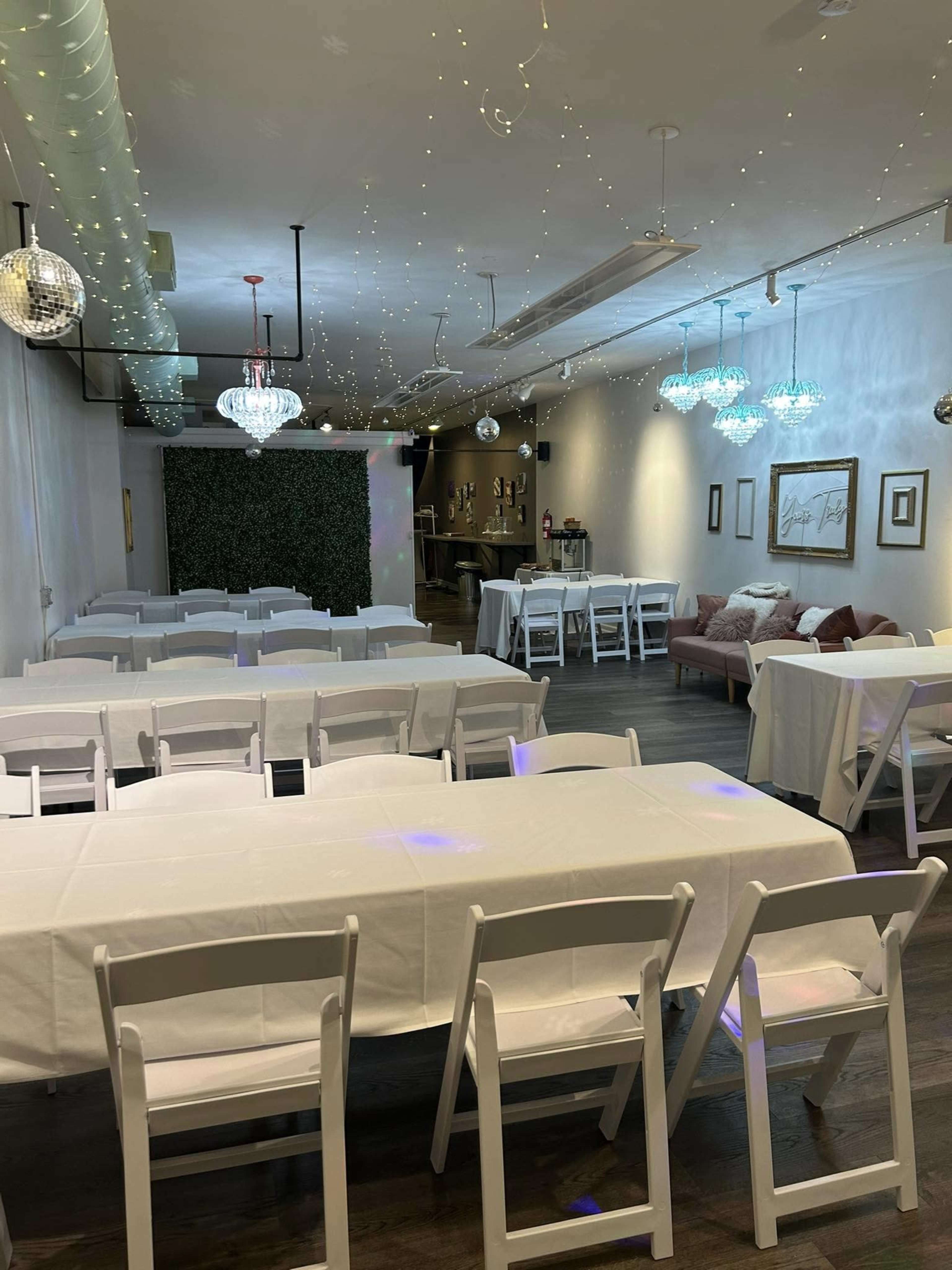 The image shows a spacious event room with white folding tables and chairs arranged neatly, twinkling lights and chandeliers hanging from the ceiling, and a small lounge area in the background.