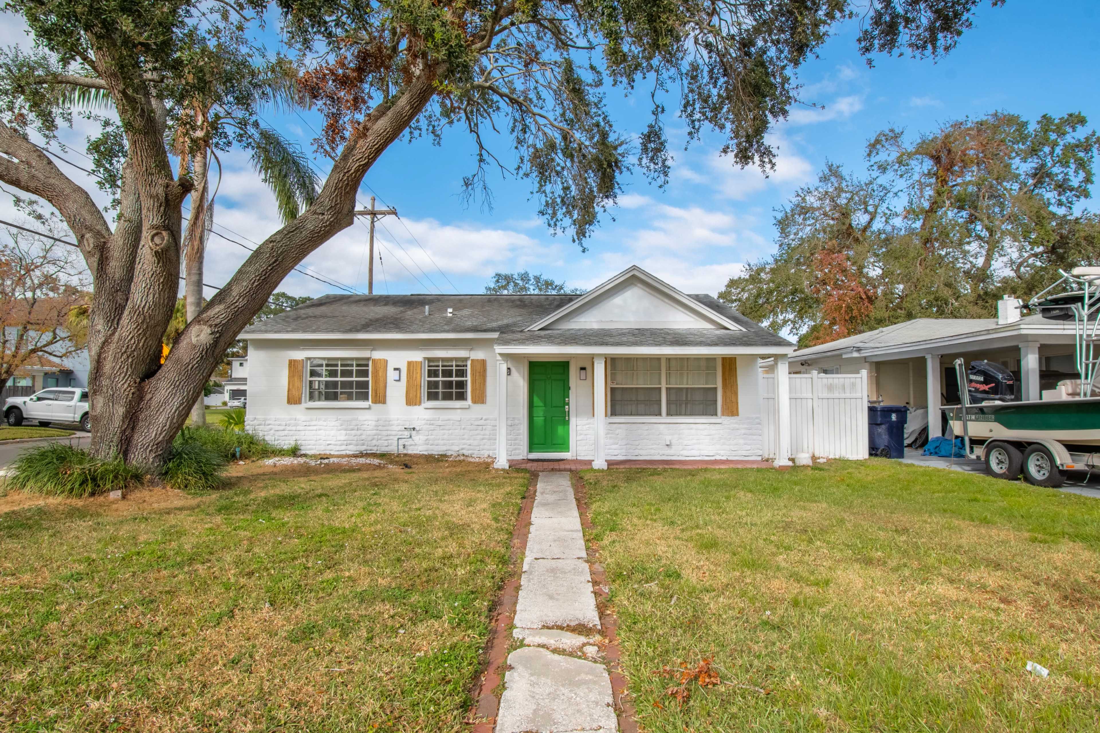 The image shows a single-story white house with green shutters and a front door, situated on a grassy lawn with a pathway leading to the entrance.