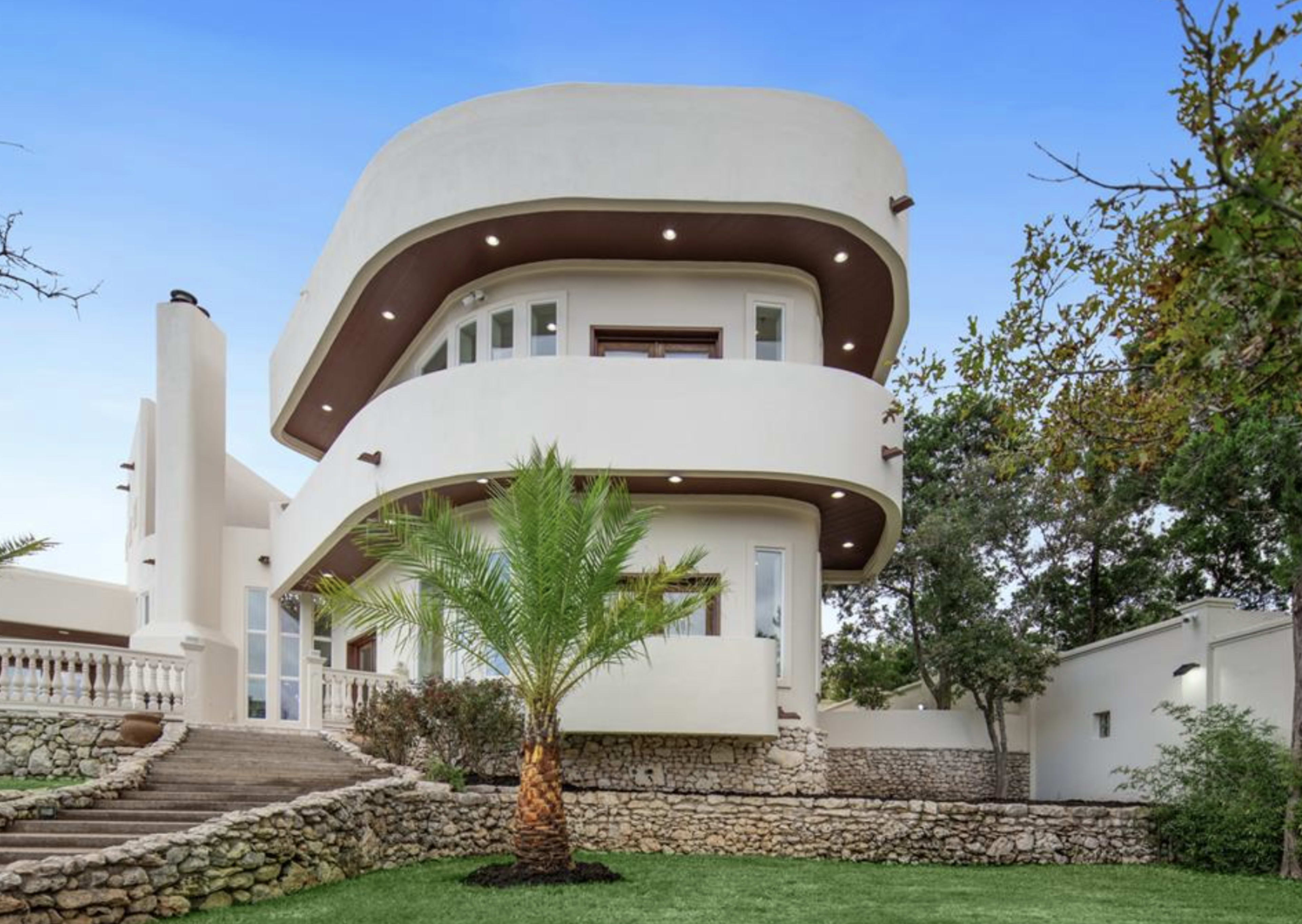 A modern two-story house with a curved architectural design, surrounded by a landscaped yard featuring a palm tree and stone retaining walls.