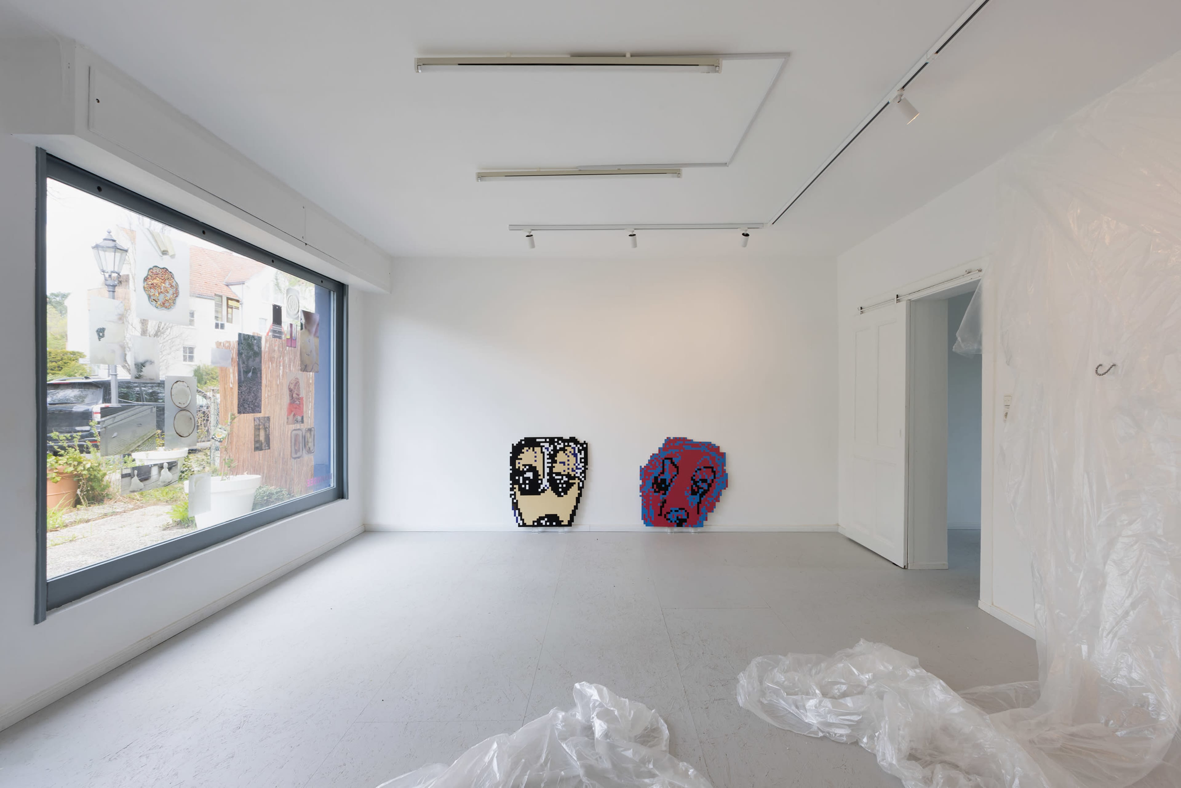 An empty gallery space with two abstract artworks featuring face-like shapes on the white walls, and plastic sheeting on the floor.