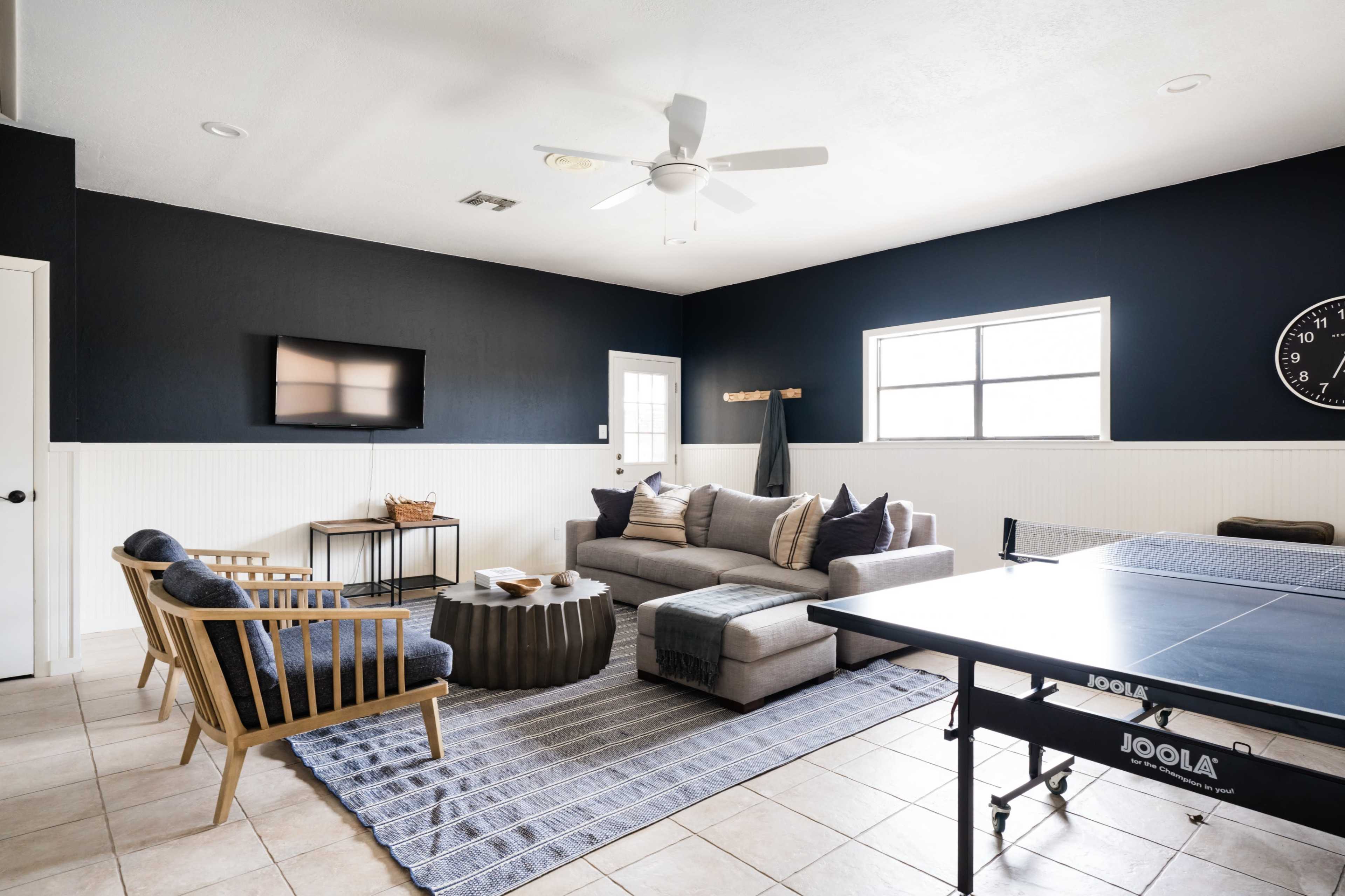 A modern living room with a gray sectional sofa, decorative pillows, a coffee table, a wall-mounted television, and a ping pong table in the corner.