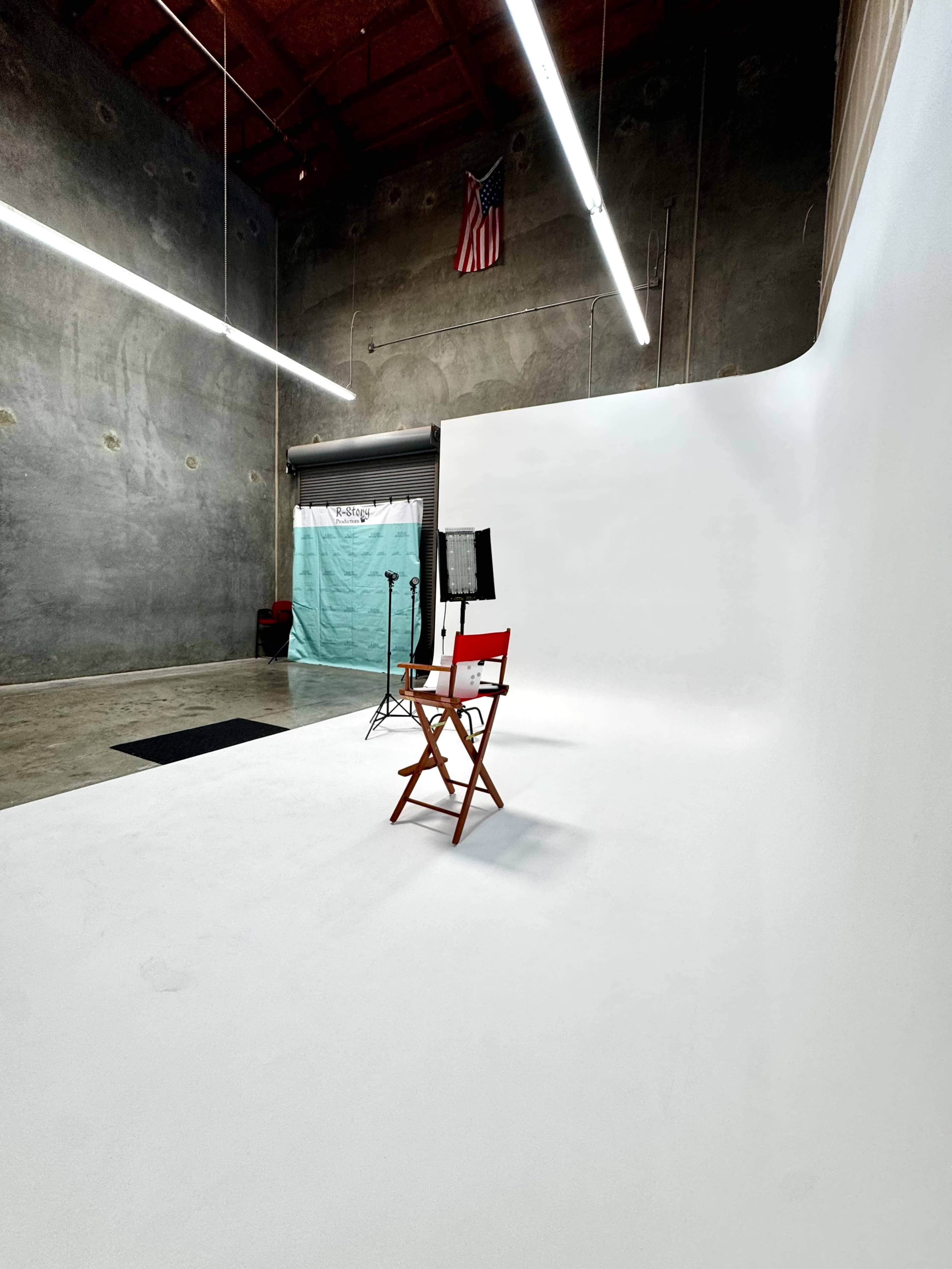The image shows a minimalist photography studio featuring a white backdrop, a director's chair, a small black mat, and a lighting setup, with the American flag visible on the wall.