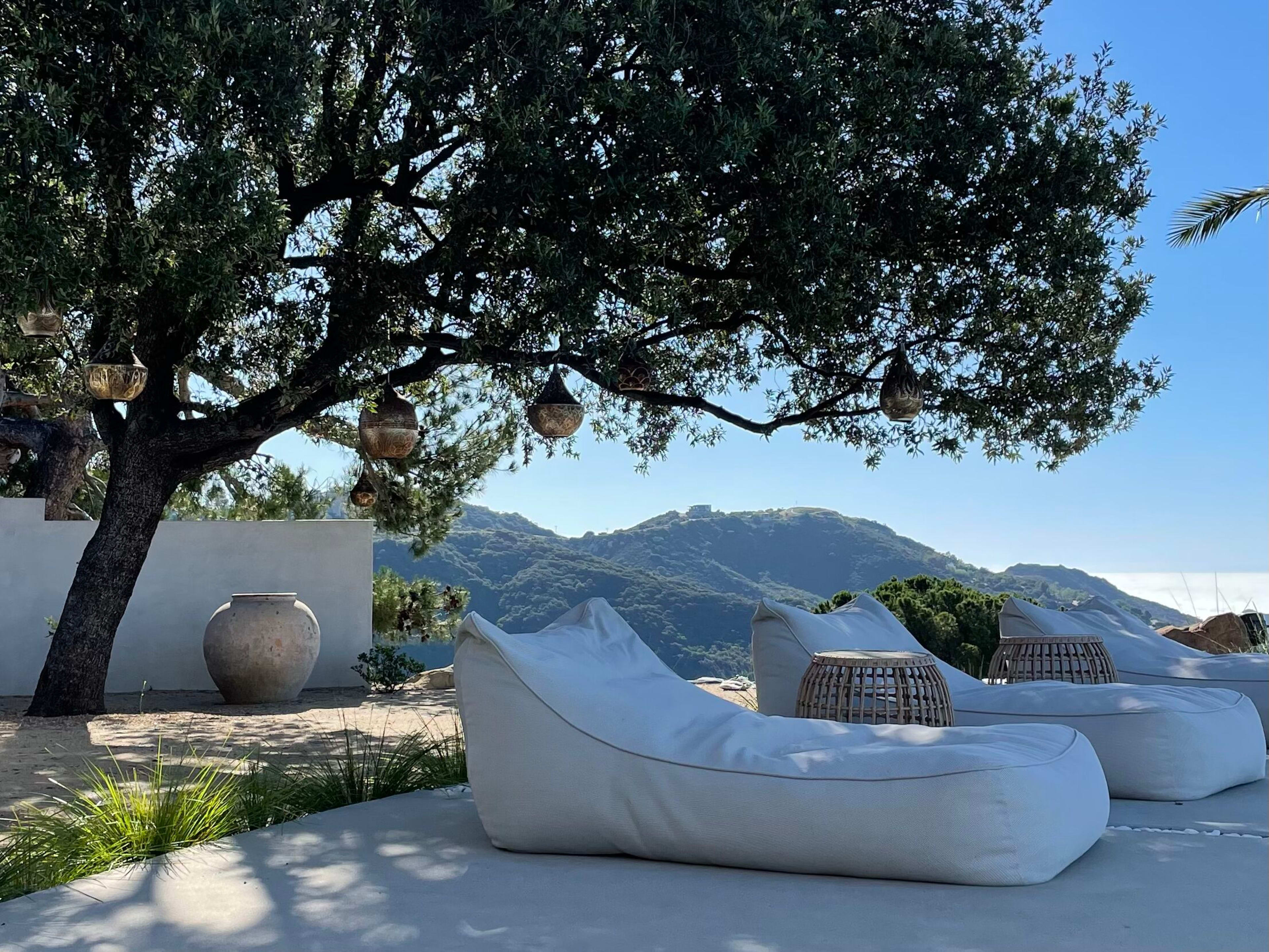 The scene features a tranquil outdoor area with white lounge cushions beneath an olive tree, overlooking green mountains in the background.