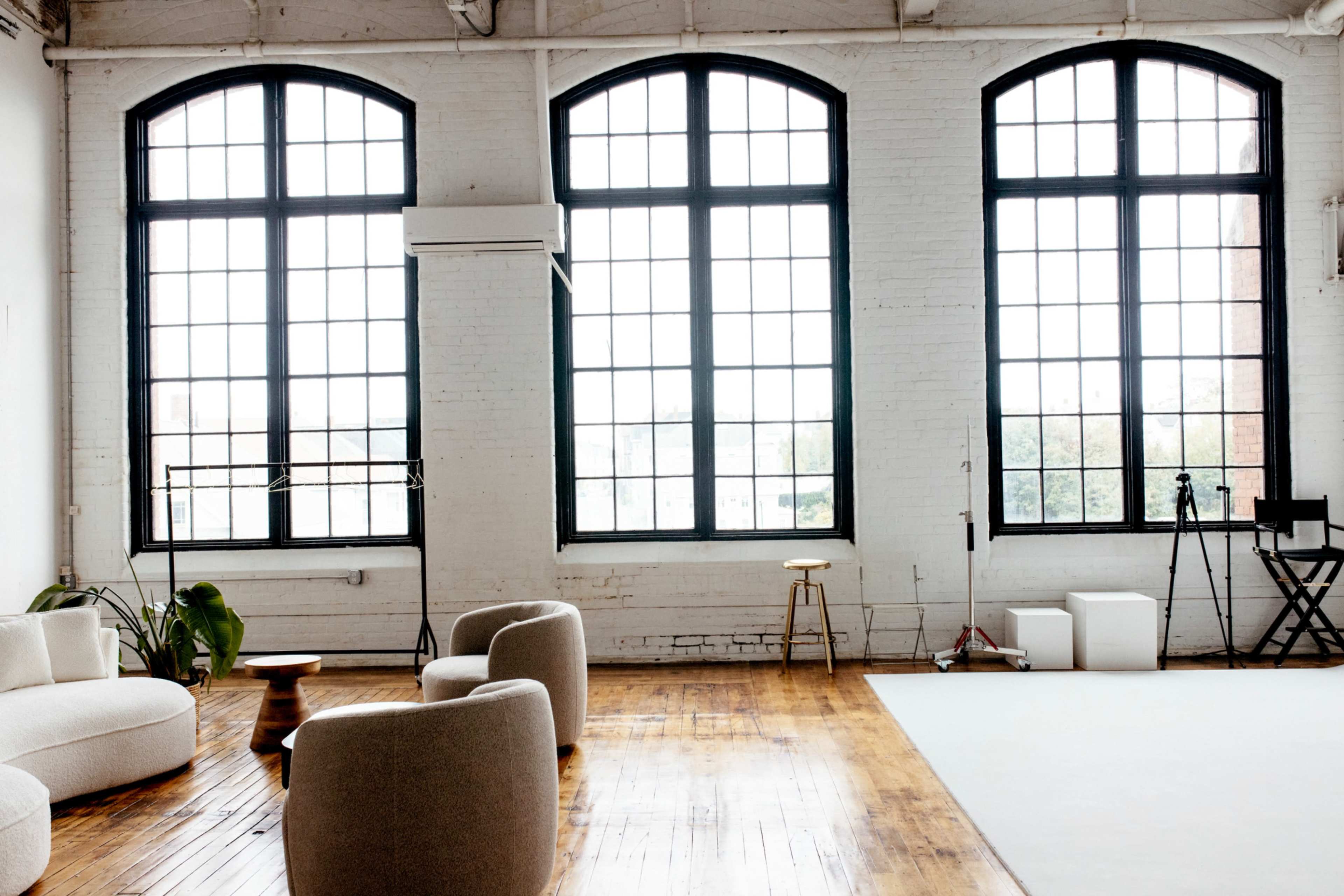 The image shows a spacious room with large arched windows, wooden flooring, and minimal furniture, including a sofa and a stool.