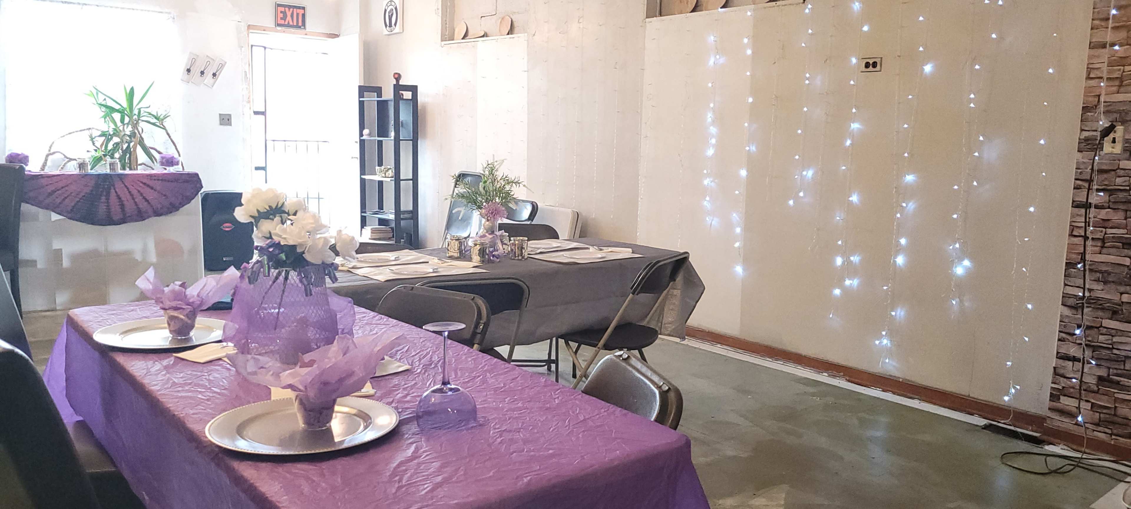 A decorated indoor space with tables set for a gathering, featuring purple tablecloths, floral arrangements, and twinkling lights on the walls.