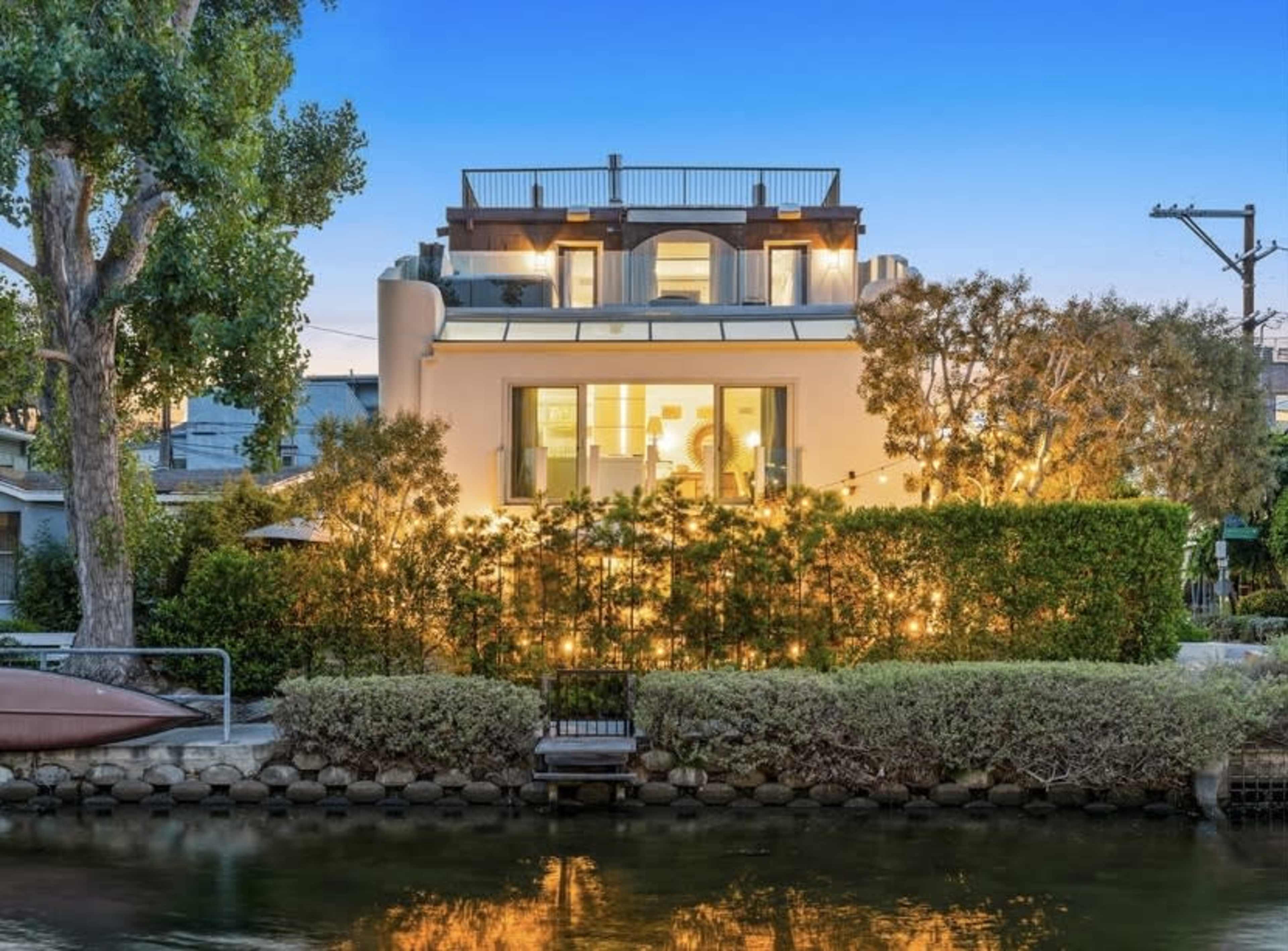A modern house with large windows sits along a canal, illuminated by exterior lights and surrounded by lush greenery.