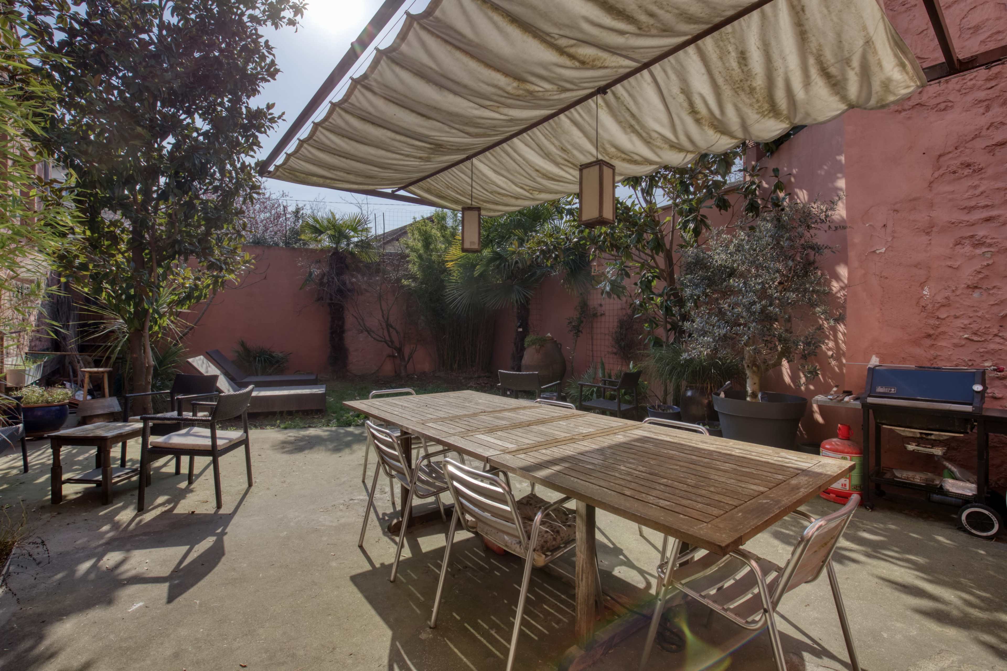 An outdoor patio area with a large wooden table, metallic chairs, a grill, and various plants surrounded by pink walls.