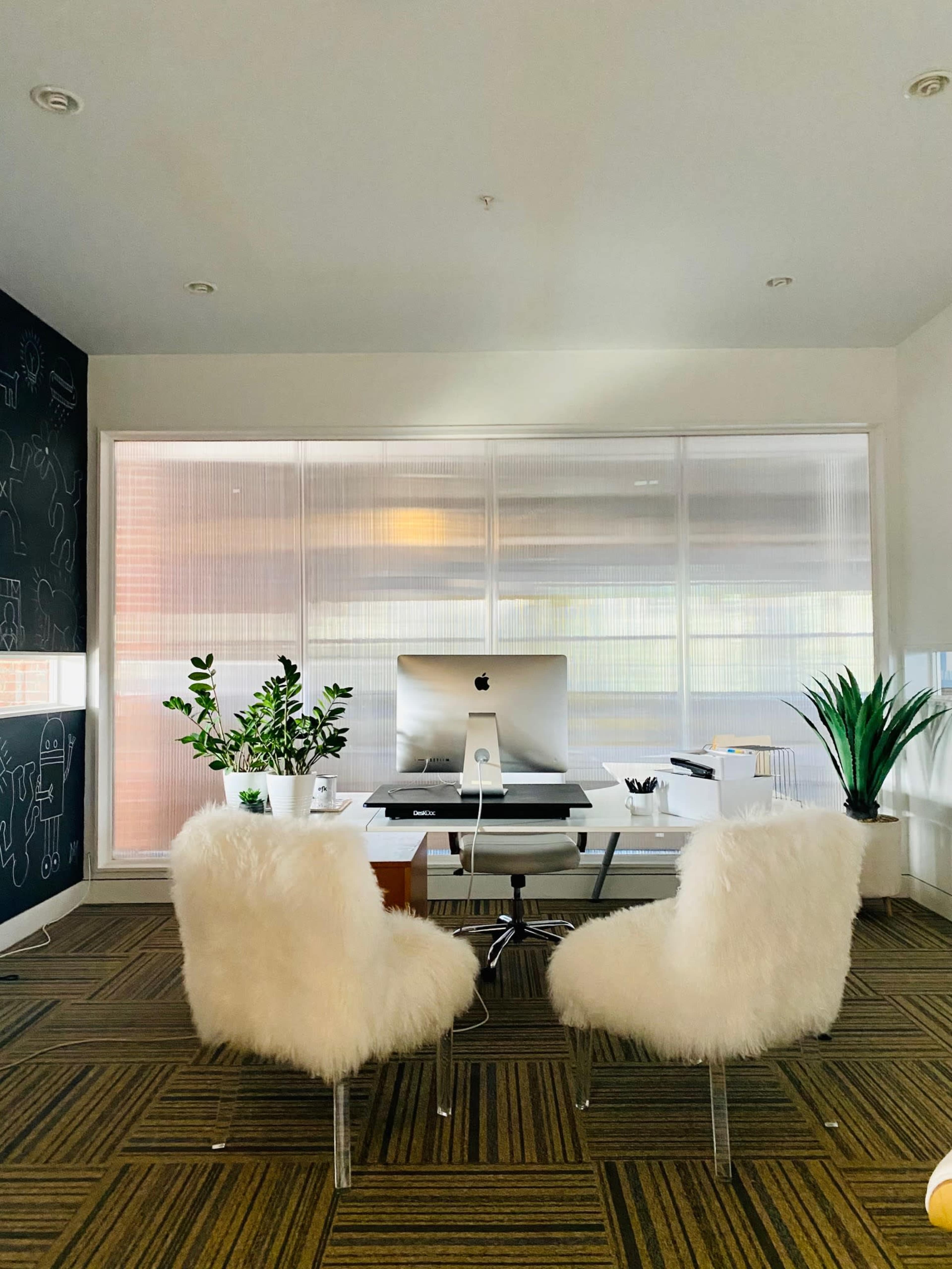 A modern office space features a white desk with an iMac, two fluffy chairs, and potted plants beside large windows.