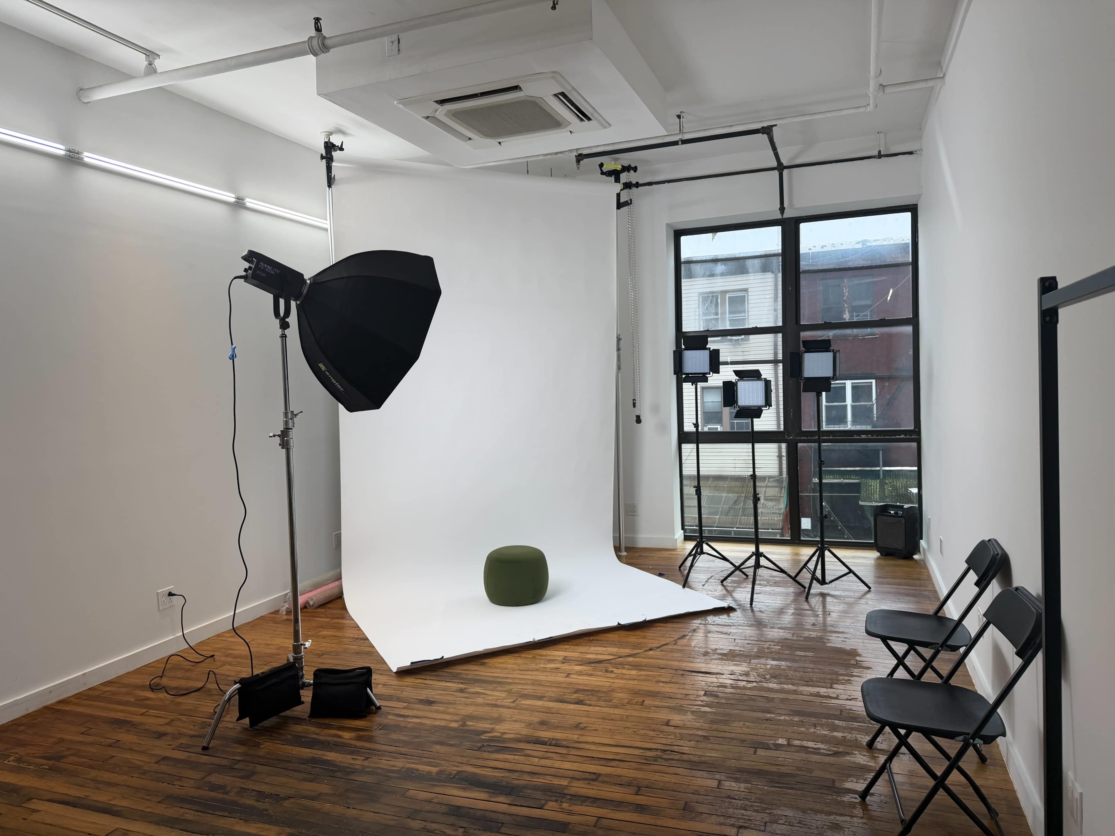 The image shows a photography studio with a white backdrop, lighting equipment, a green stool, and several black folding chairs arranged nearby.