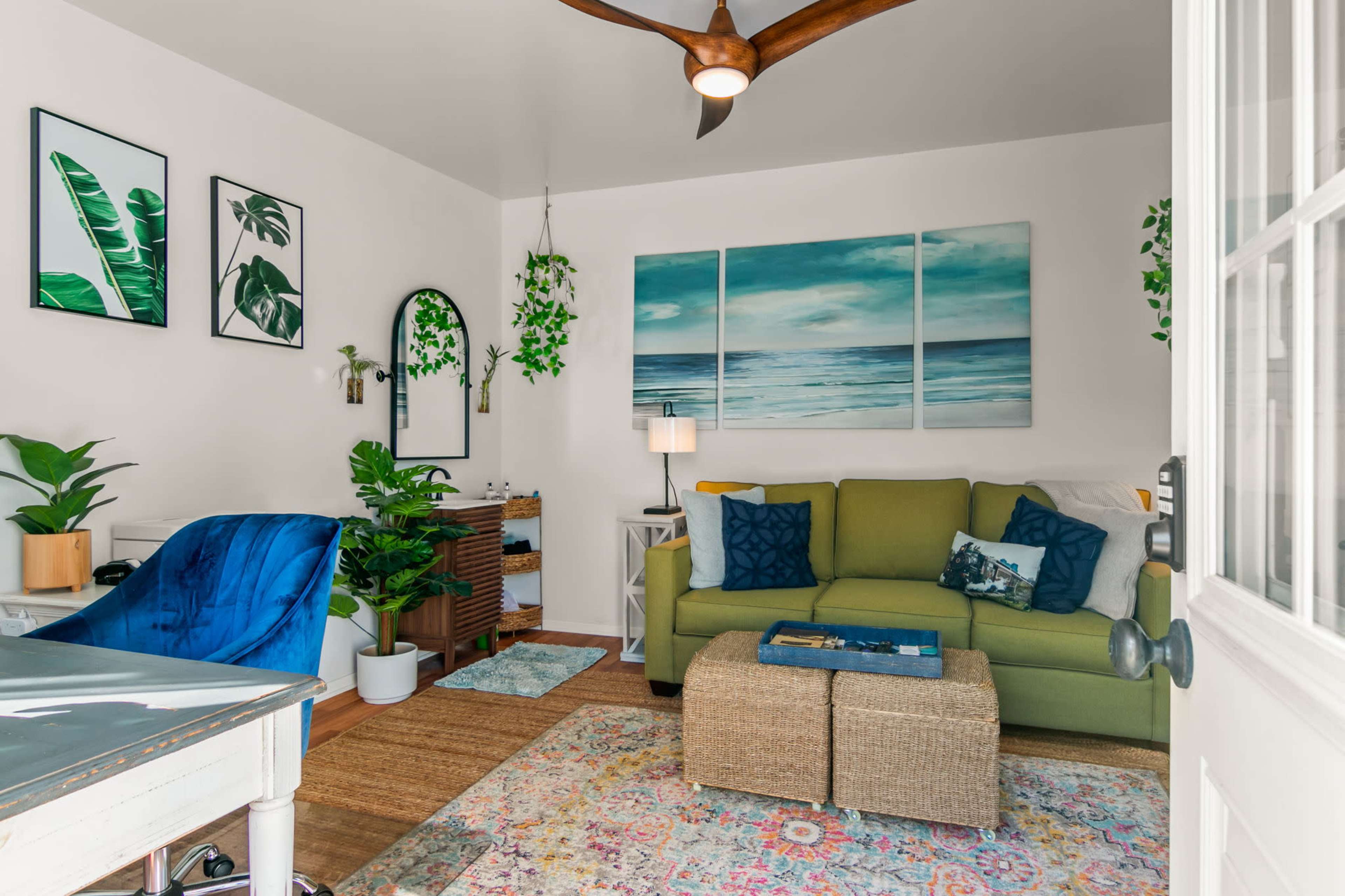 A bright and stylish living room features a green sofa, a wooden desk, a large rug, and framed botanical artwork on the walls.