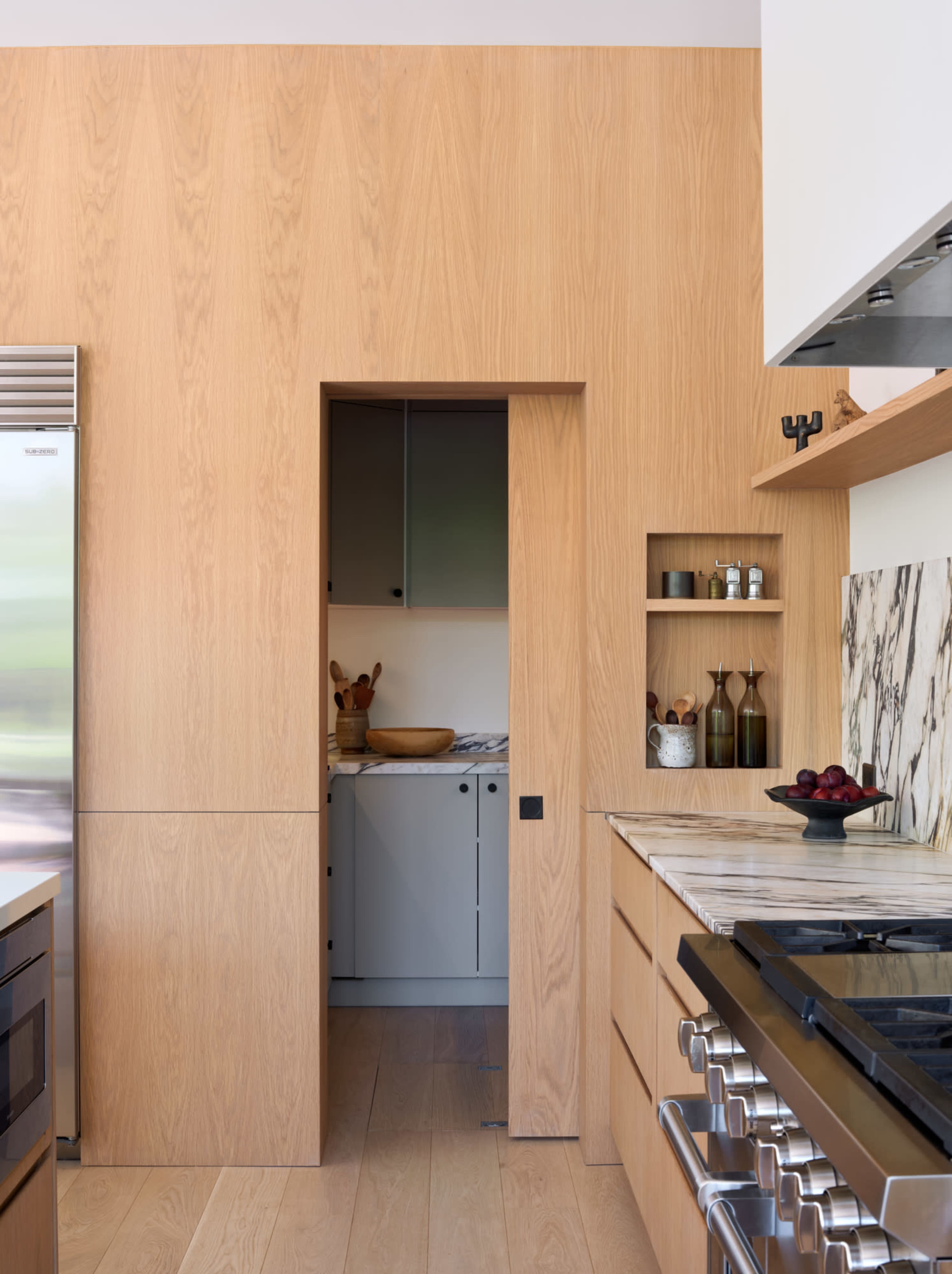 The image shows a modern kitchen featuring wooden cabinets, a marble countertop, and a small open doorway leading to a pantry or utility area.