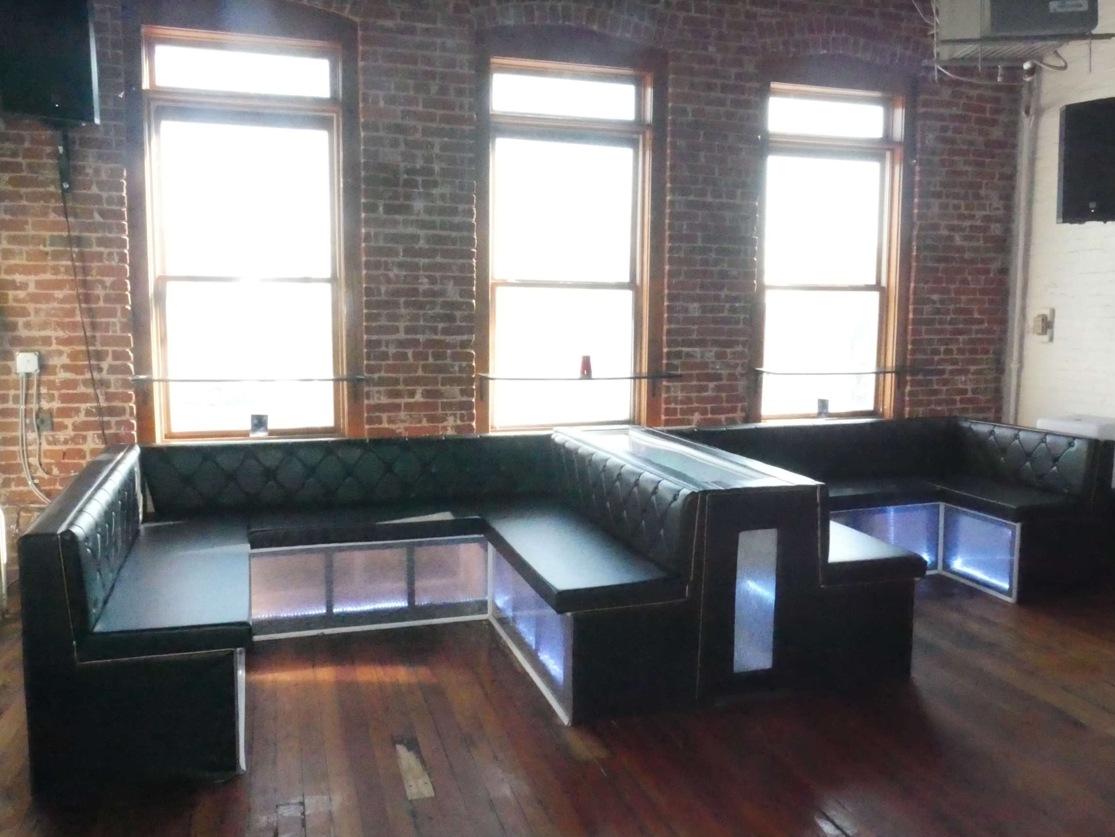 A spacious lounge area with black leather benches and illuminated sections, set against a backdrop of exposed brick walls and large windows.
