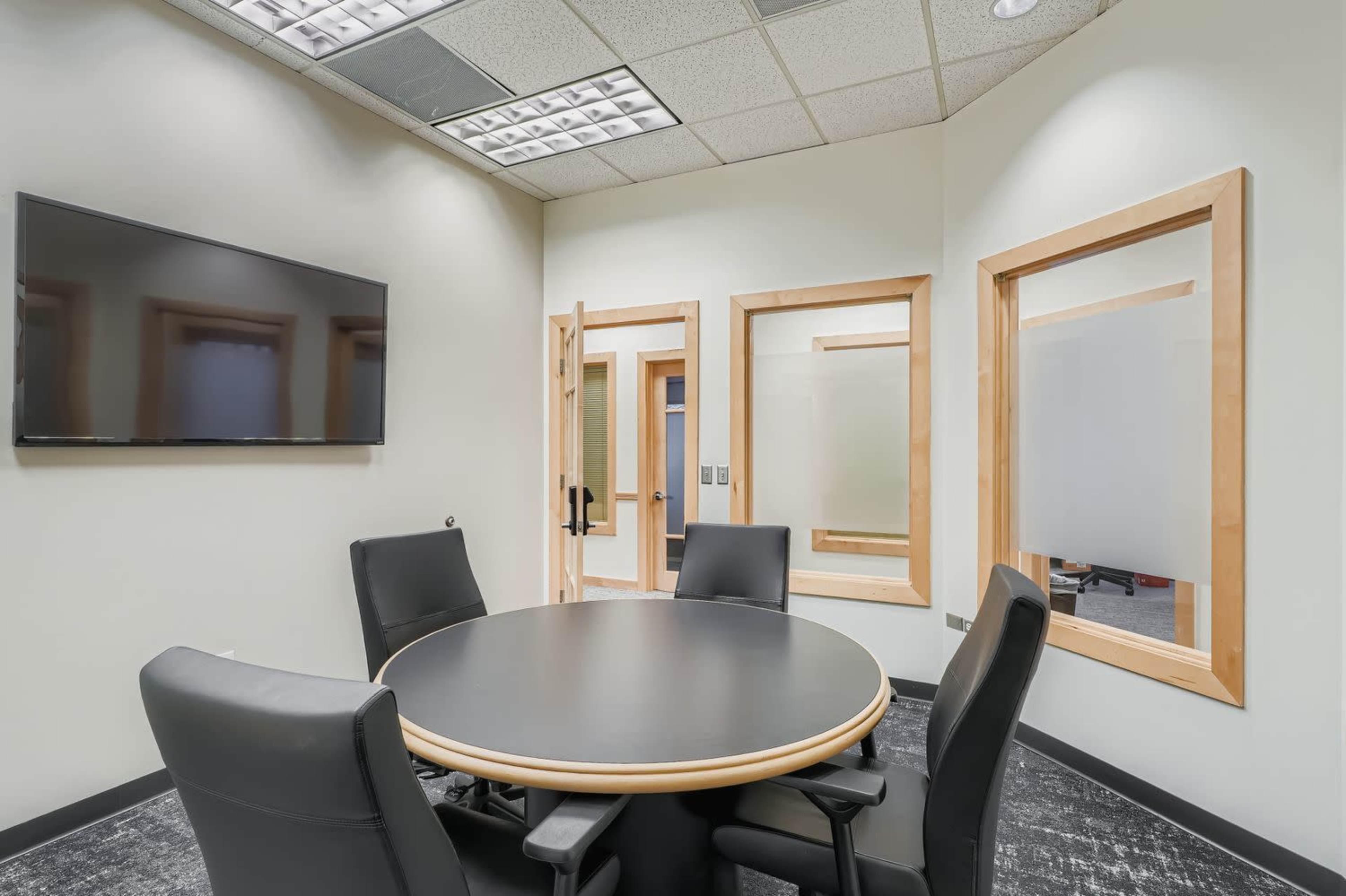The image shows a conference room featuring a circular table surrounded by four black chairs, a large wall-mounted television, and glass panel doors.