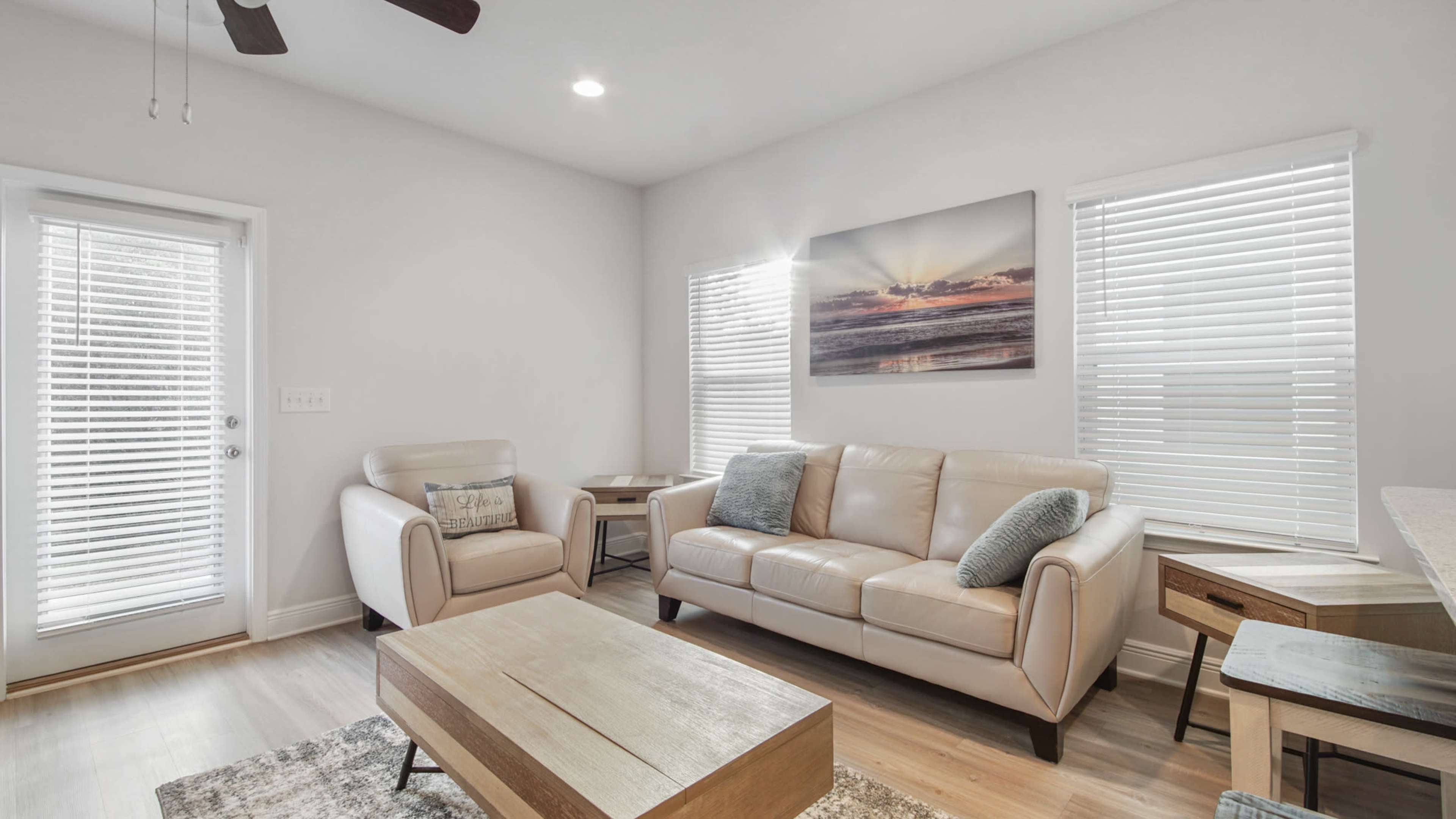 The image shows a bright, modern living room featuring a light-colored leather sofa, a wooden coffee table, and large windows with blinds.